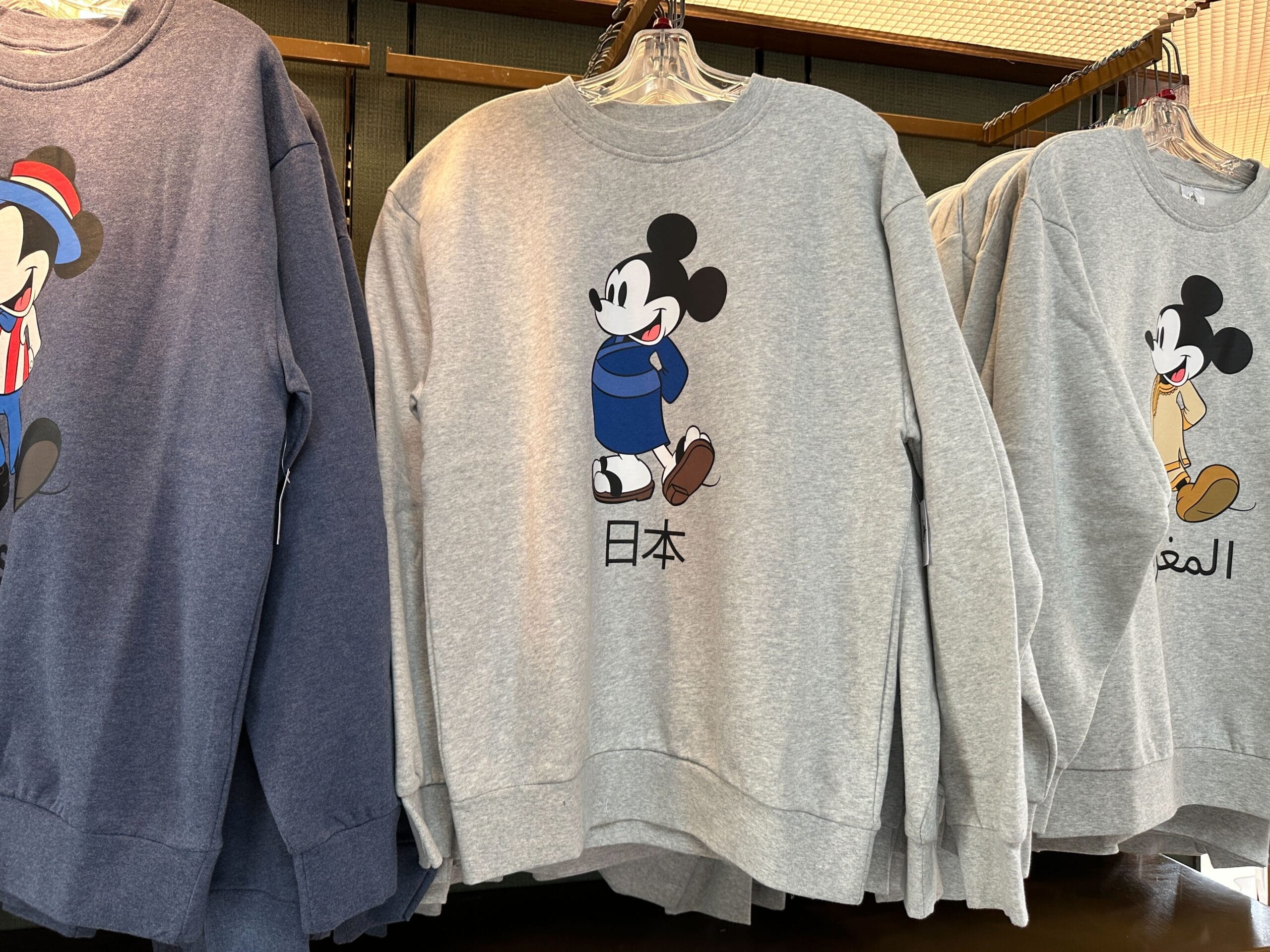 Gray World Showcase Sweatshirt featuring Mickey Mouse in blue clothes and Japanese characters, hanging on a rack with others.