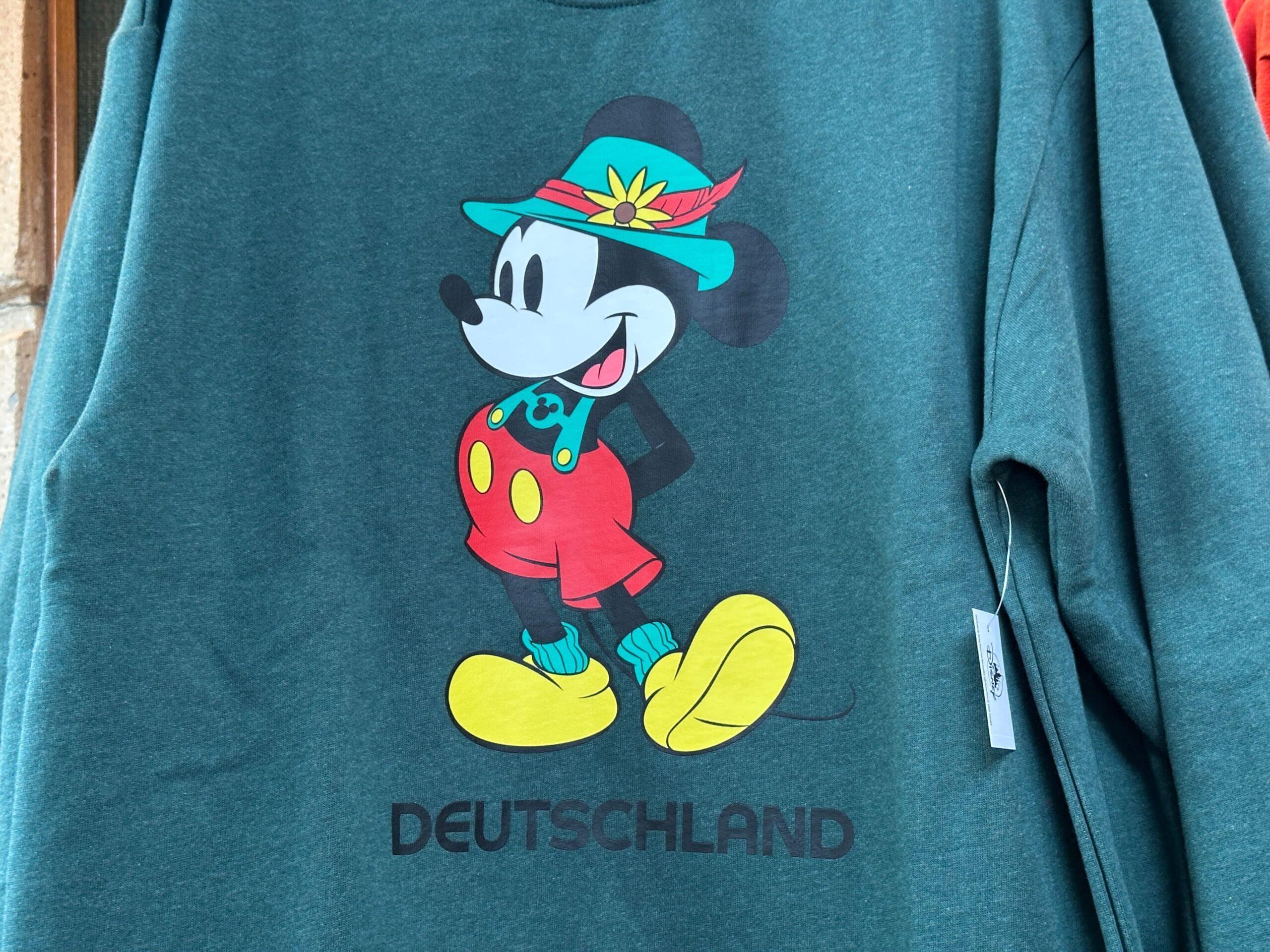 Mickey Mouse in Bavarian attire with "DEUTSCHLAND" below, part of the EPCOT Sweatshirt Collection on a green shirt.