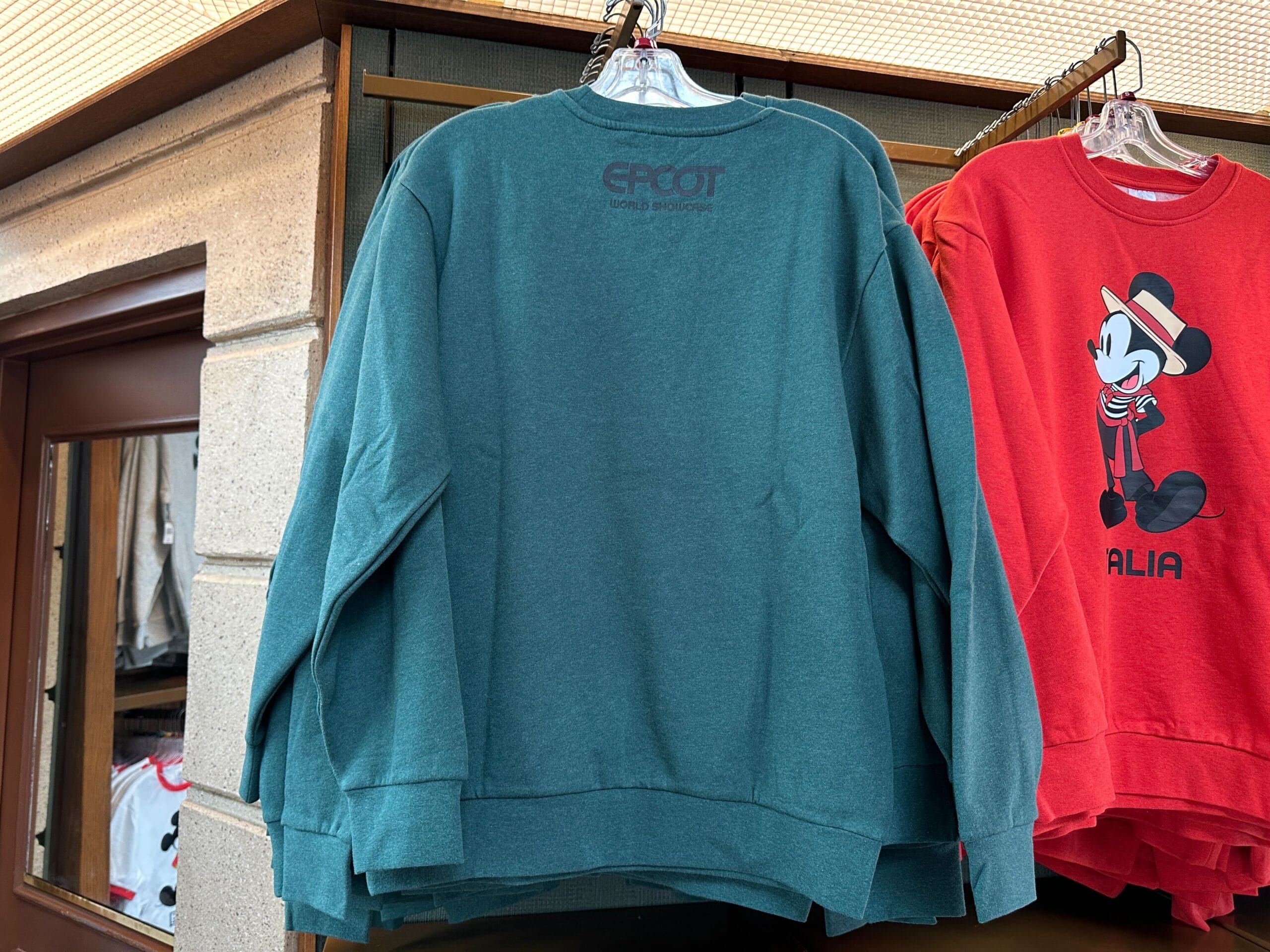 Green "EPCOT" sweatshirt from the Sweatshirt Collection hangs beside a red Mickey Mouse graphic sweatshirt in-store.
