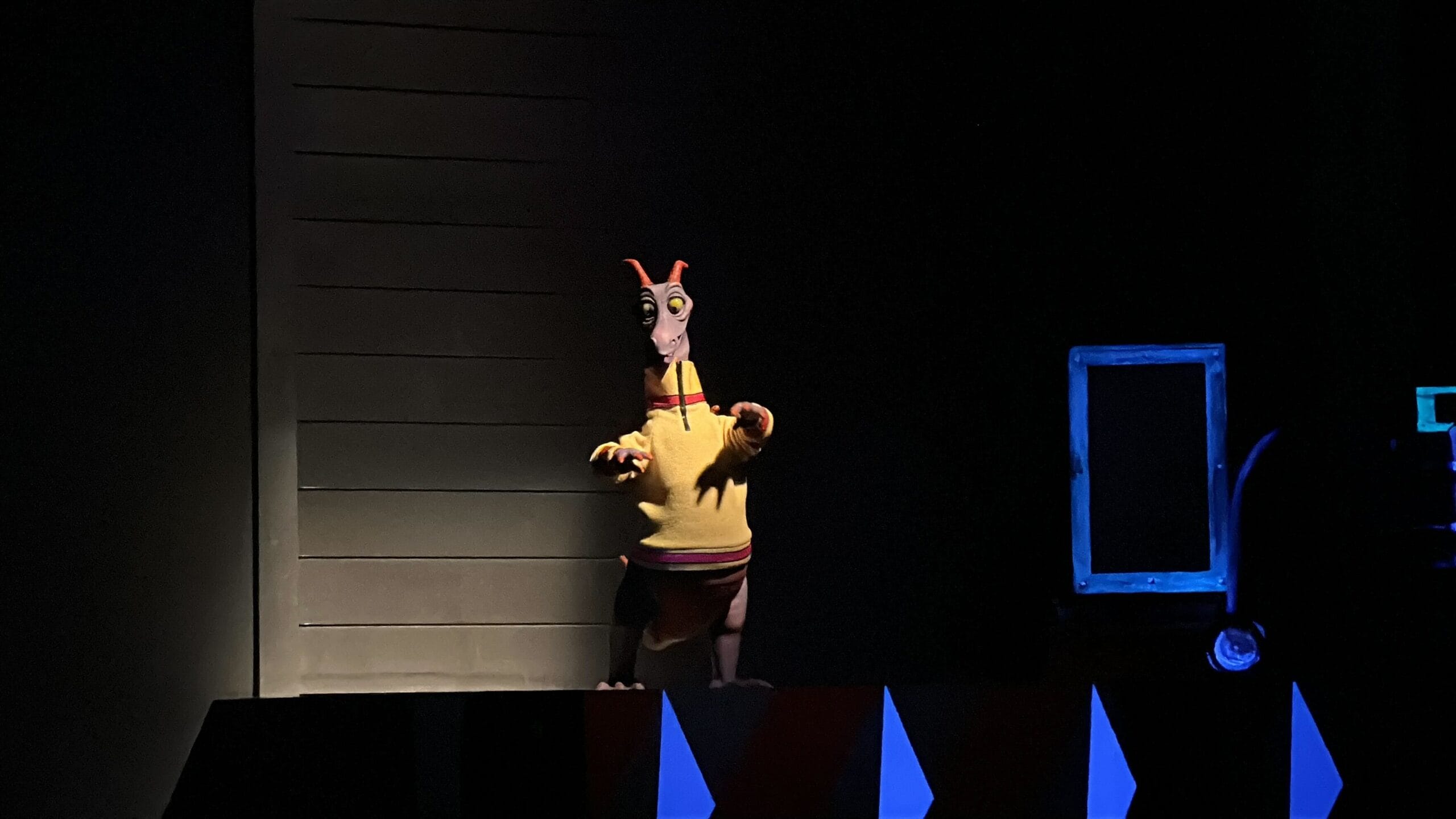 A Figment animatronic in a yellow shirt stands illuminated on a dark Disney park stage next to a blue-framed rectangular portal.