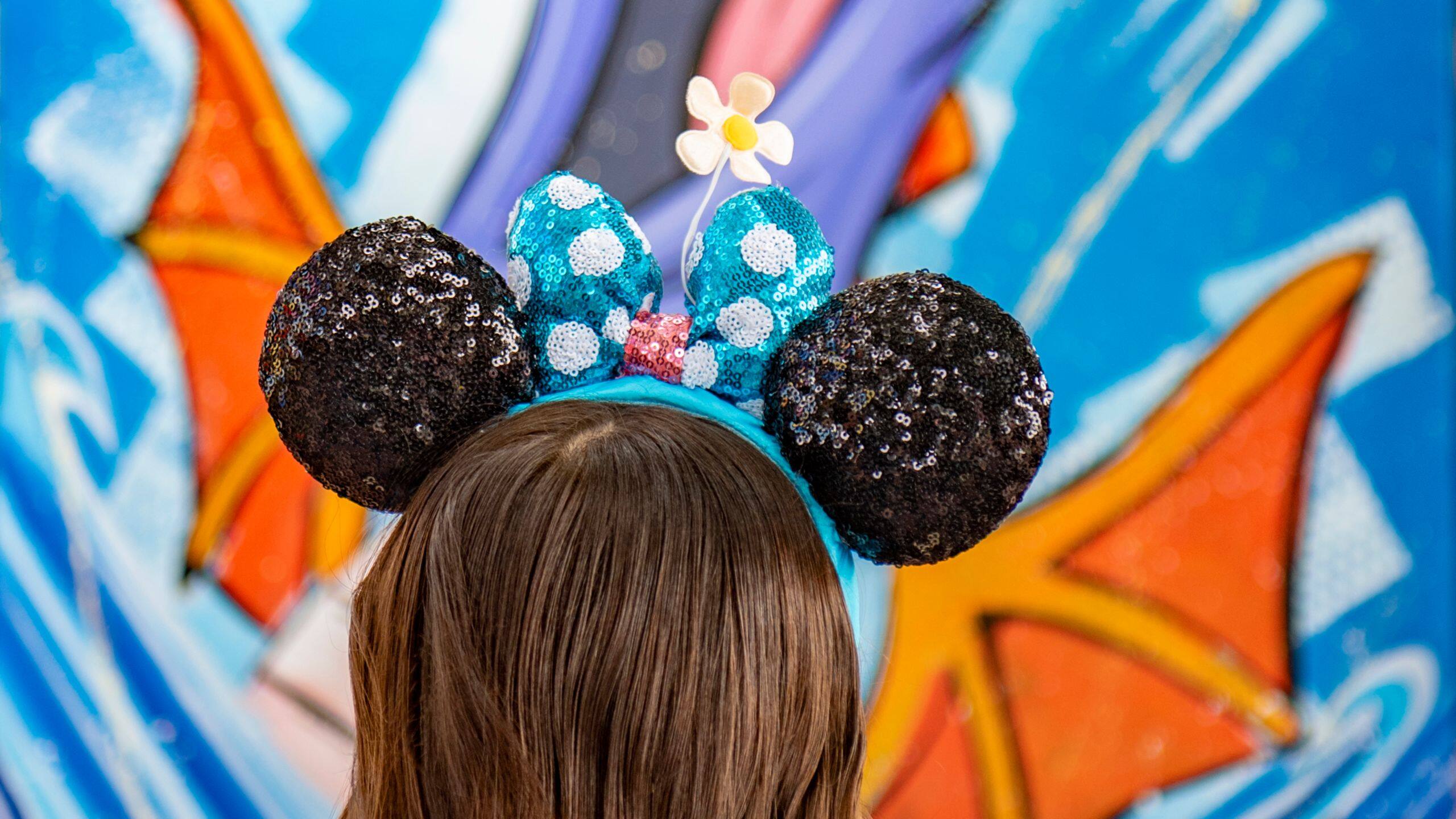 A guest in sparkling Minnie Mouse ears poses by a vibrant mural at EPCOT Festival of the Arts 2026, with no castle in view.