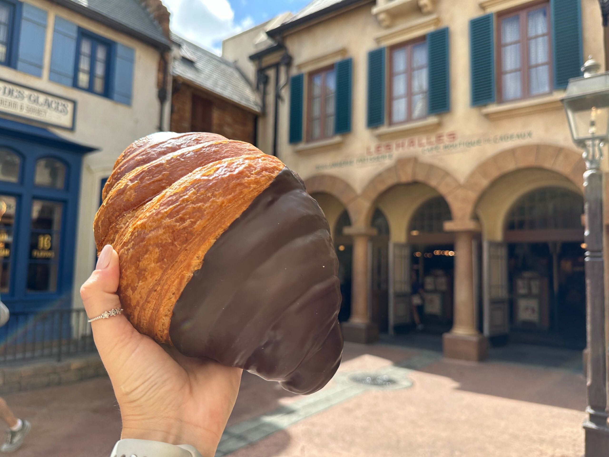 A hand holds a Giant Chocolate Twist Croissant in front of the EPCOT French bakery for a mouthwatering review.