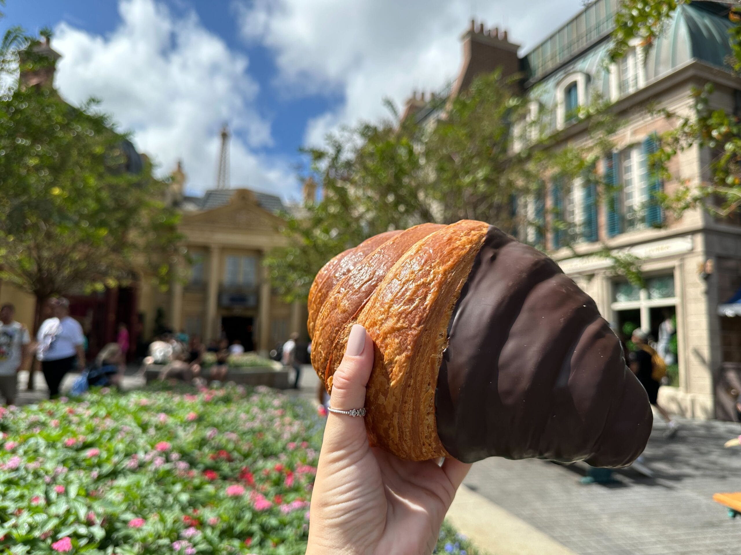 A hand holds a Giant Chocolate Twist Croissant in a sunny, garden-filled spot near European-style buildings at EPCOT.