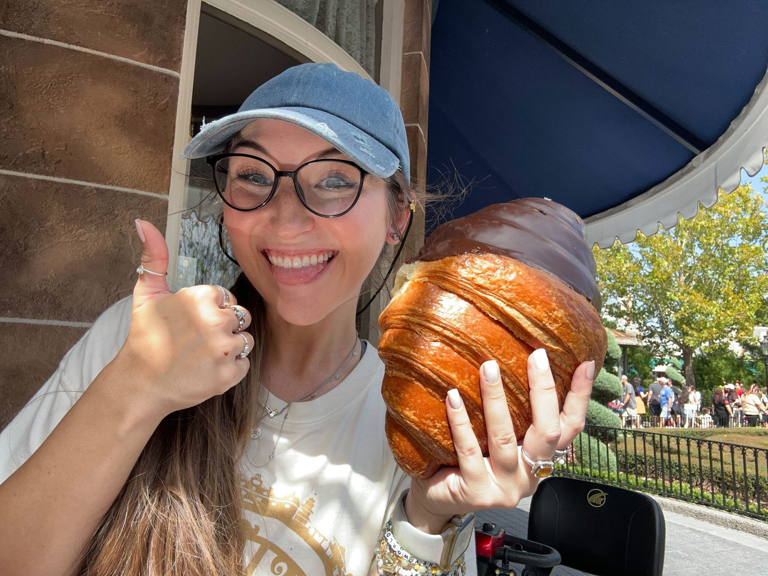 Smiling woman in glasses reviews a Giant Chocolate Twist Croissant and gives a thumbs up outside under a blue EPCOT awning.