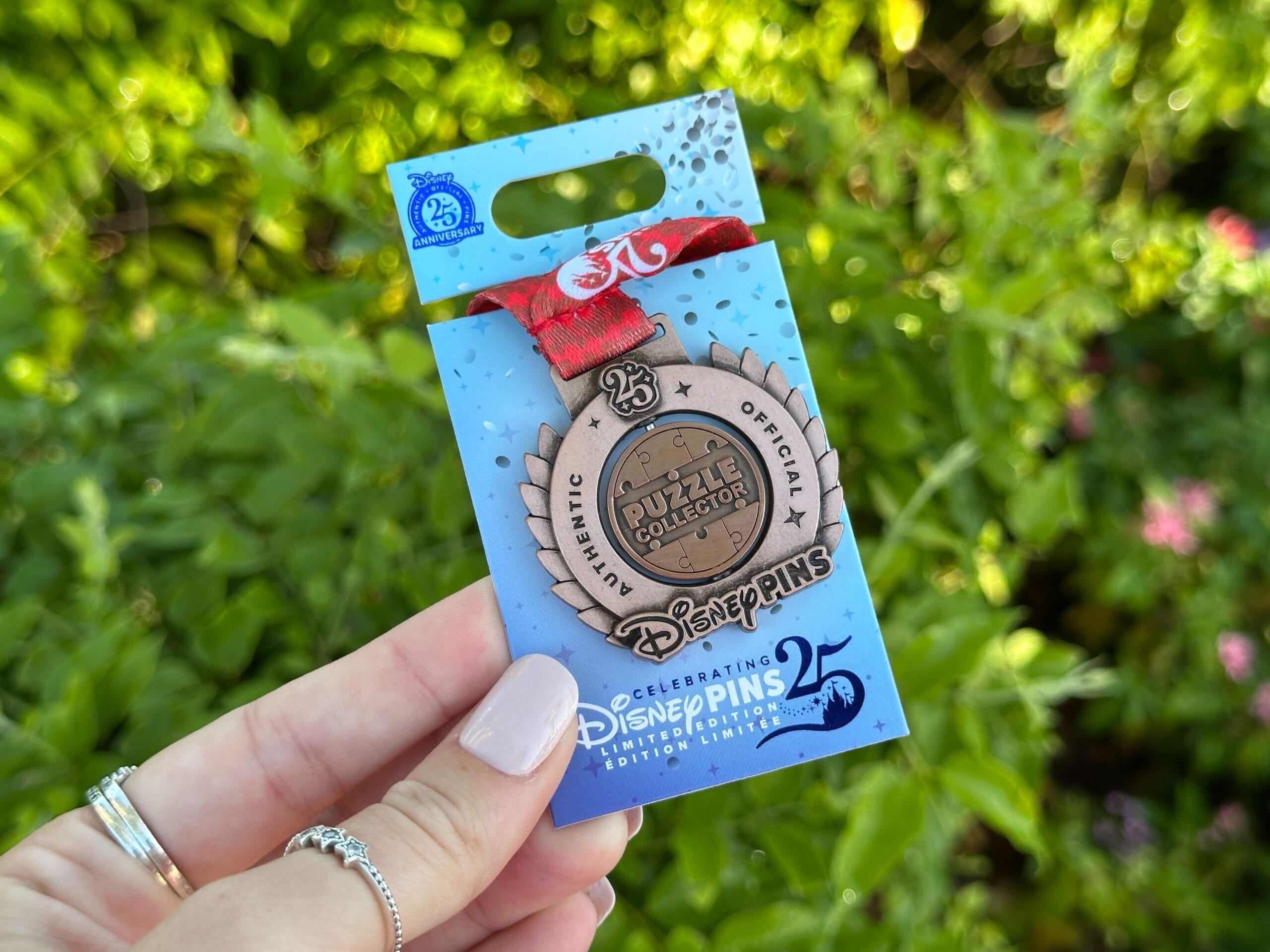 A hand holds a Disney World 25th anniversary medal pin on a blue card; lush greenery behind, no castle like Cinderella Castle visible.