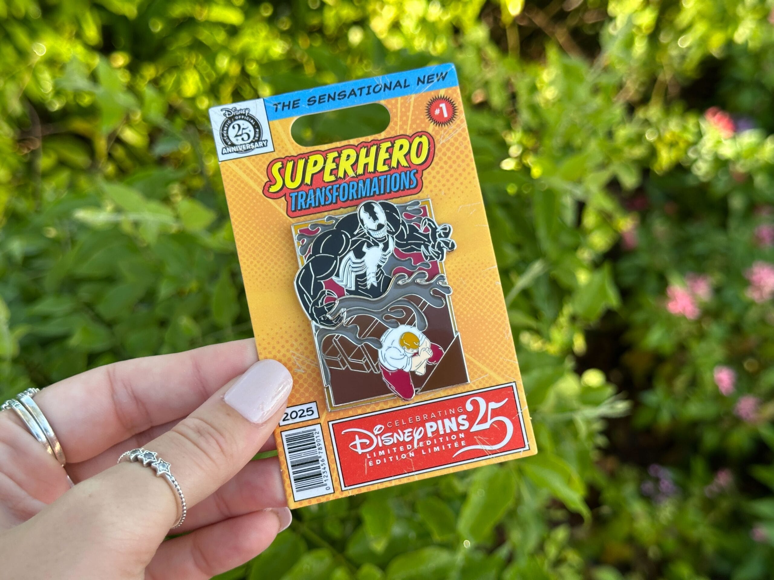 A hand displays a Disney World Pin collectible in its packaging, set against lush green foliage, evoking the magic of the parks.