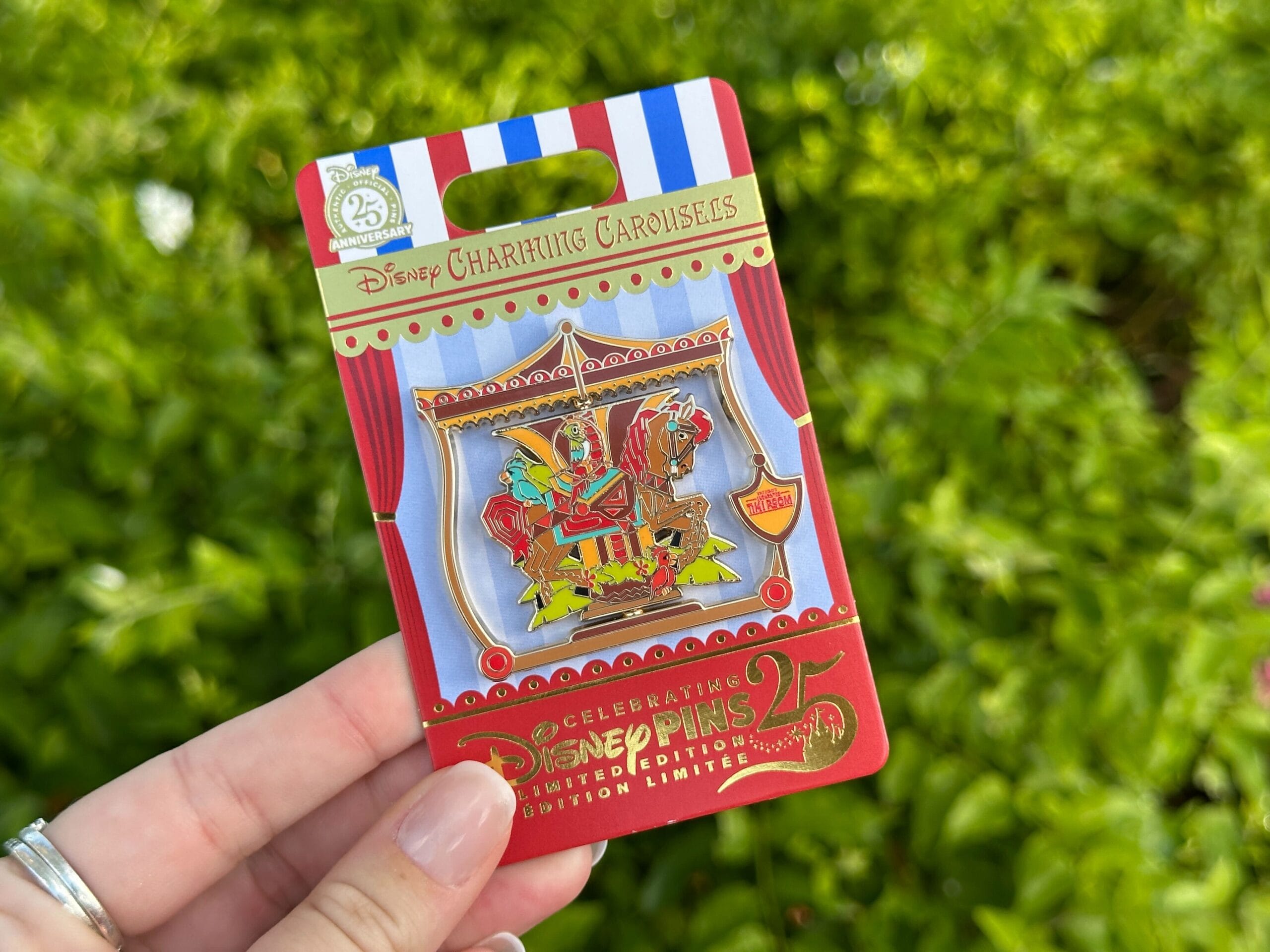A hand holds a Disney World pin of a carousel horse, with leafy greenery behind