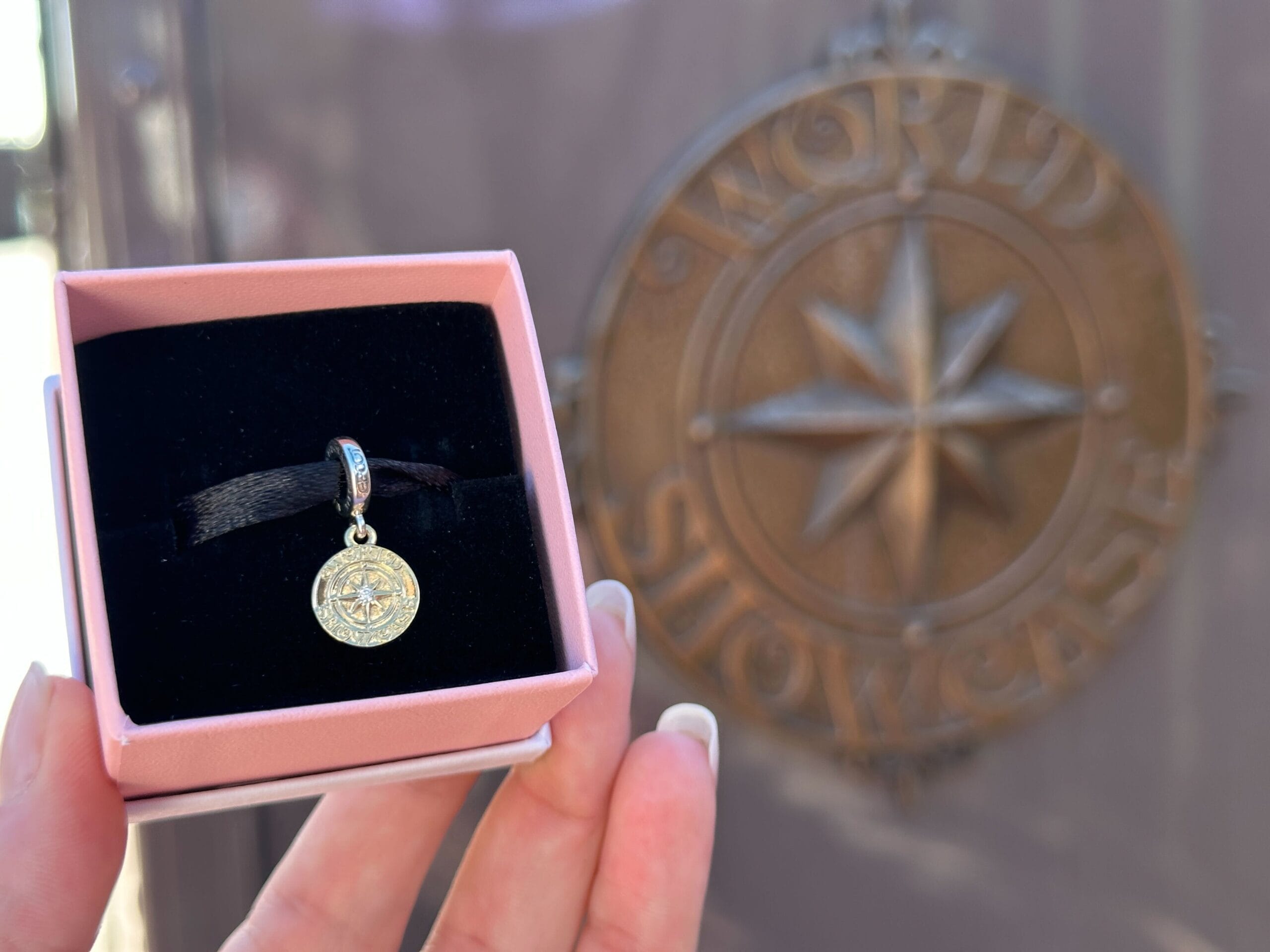 A hand holds a small World Showcase compass charm in a box, reminiscent of Epcot at Walt Disney World, bronze emblem blurred behind.