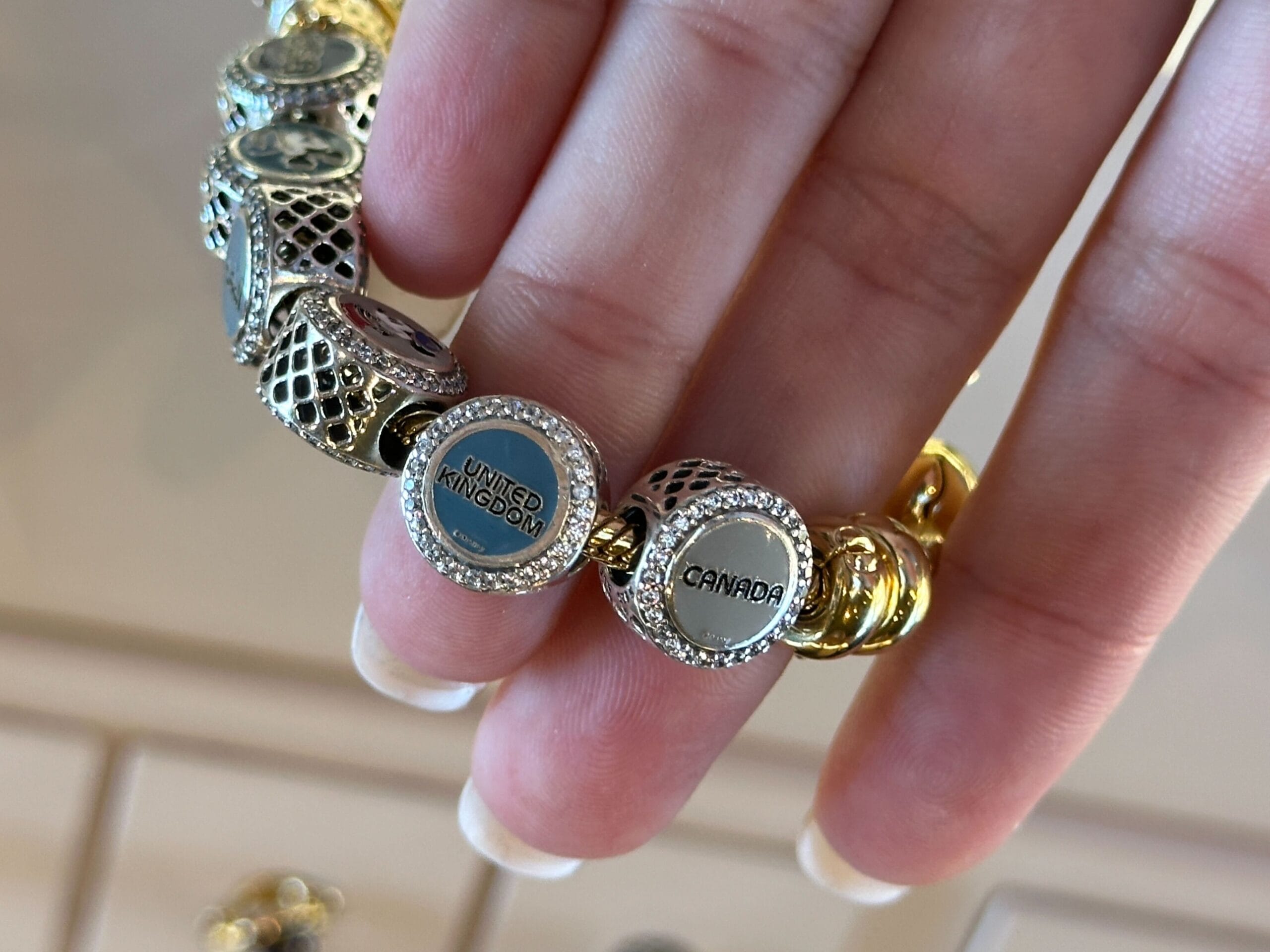 A hand holds a World Showcase Charms bracelet featuring "UNITED KINGDOM" and "CANADA" beads, a nod to EPCOT’s global pavilion flair.