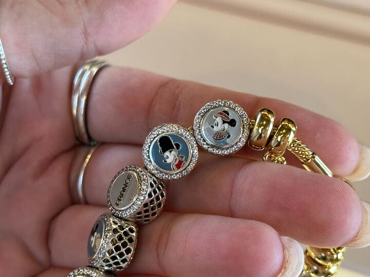 A hand displays a bracelet with World Showcase Charms and beads, plus a Mickey Mouse in a black hat—Disney Parks collectors’ style.