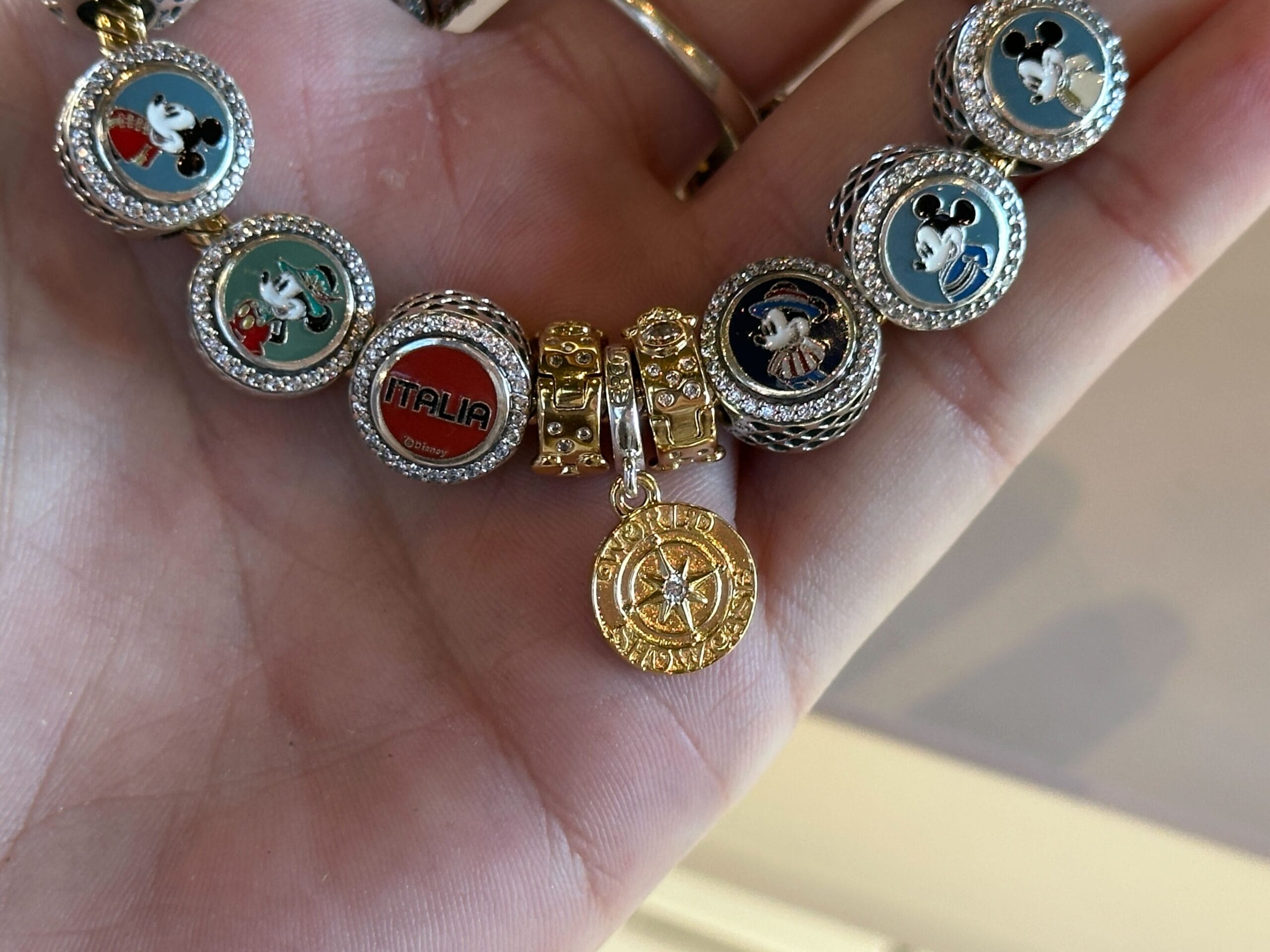 A hand displays a bracelet featuring Mickey, Minnie, and World Showcase charms, plus a gold Italia medallion like at EPCOT.