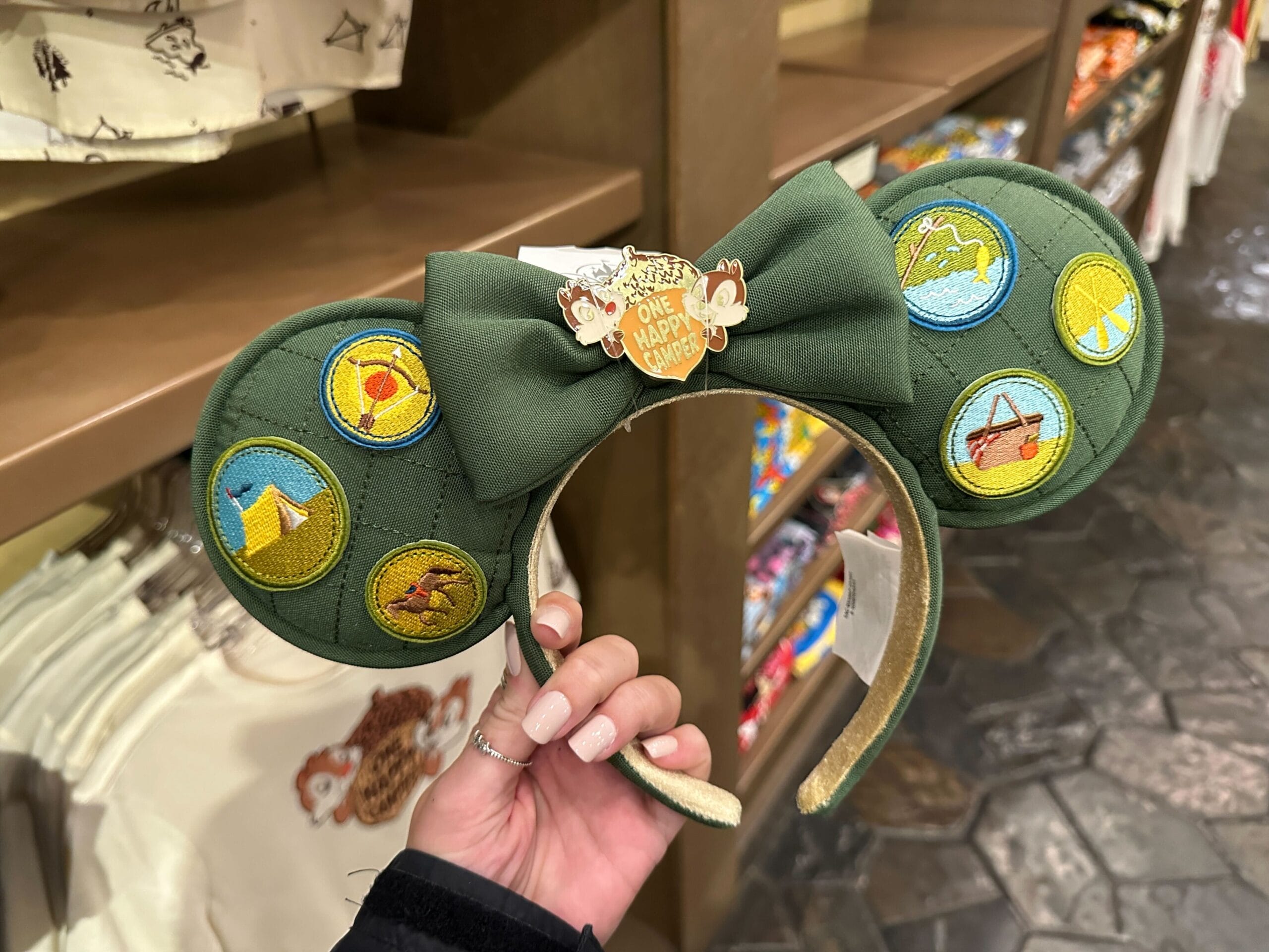 A hand holds a green Disney Ear Headband with camping patches and a "One Happy Camper" badge at Fort Wilderness display.