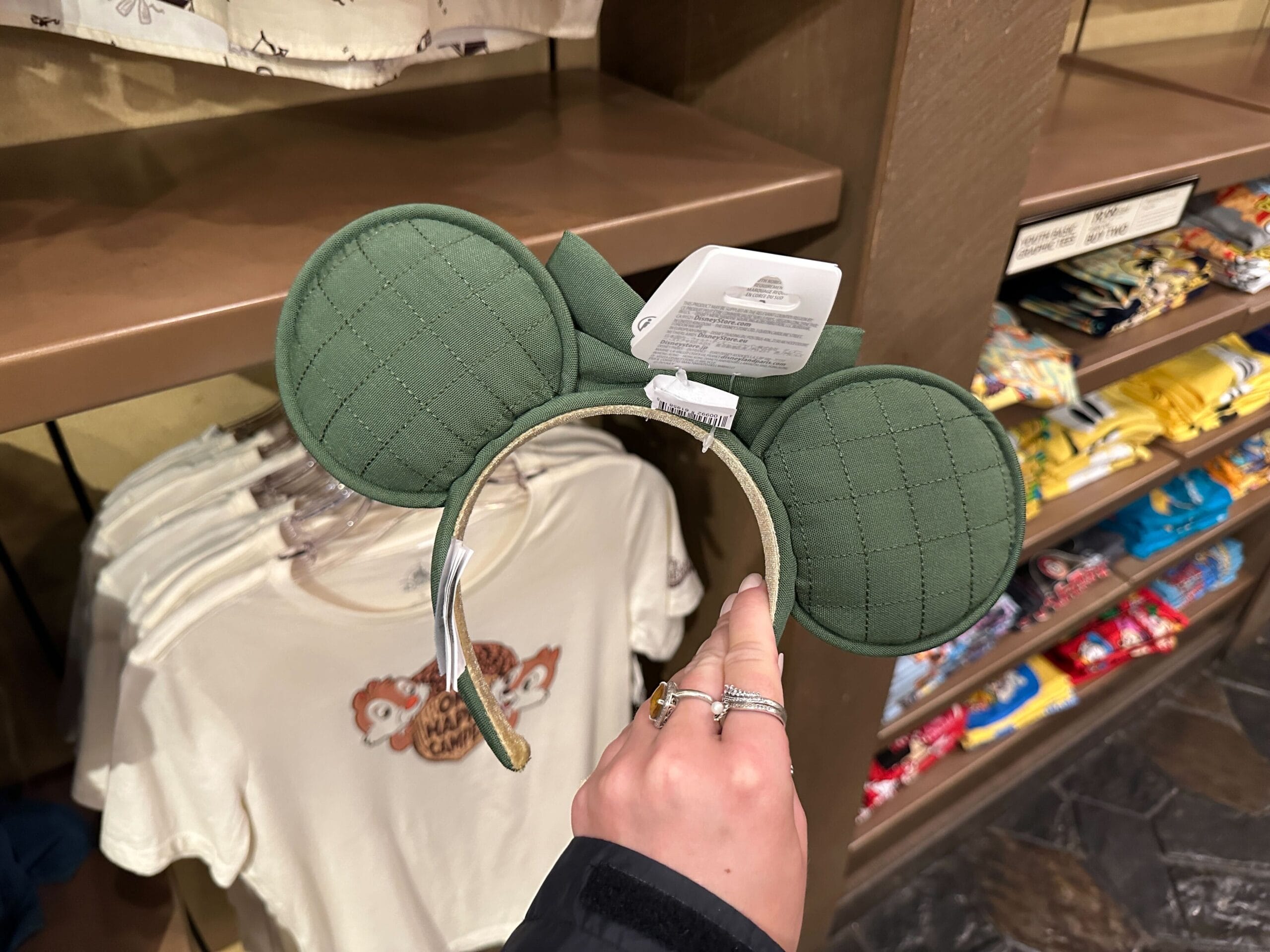 A hand holds a green quilted Disney Ear Headband in front of Fort Wilderness shirts and merchandise.
