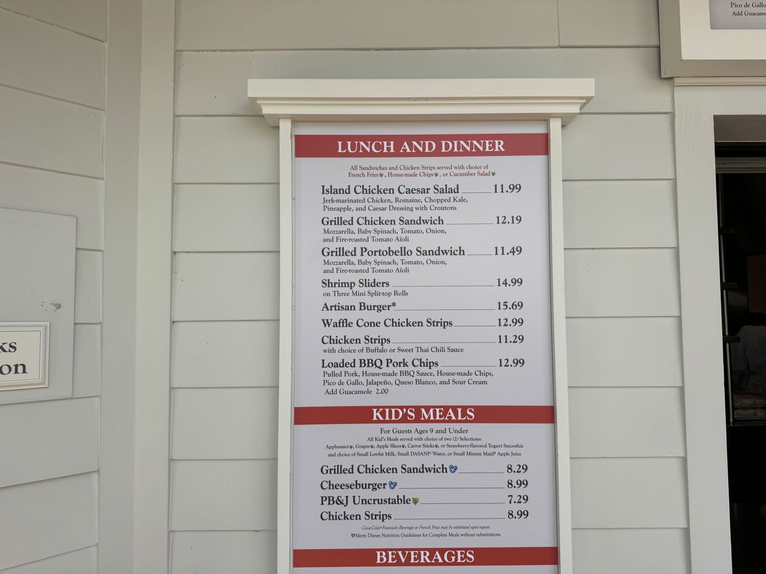 Menu board at a Disney park restaurant listing lunch, dinner, chicken strips, kids' meals, and prices, mounted on the wall.