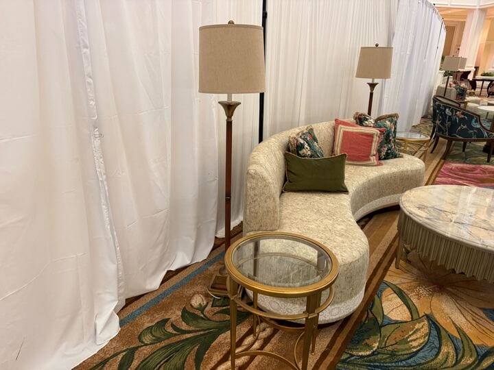 Curved sofa with assorted pillows, round glass table, and floor lamp evoke a Disney resort lounge near sheer curtains.