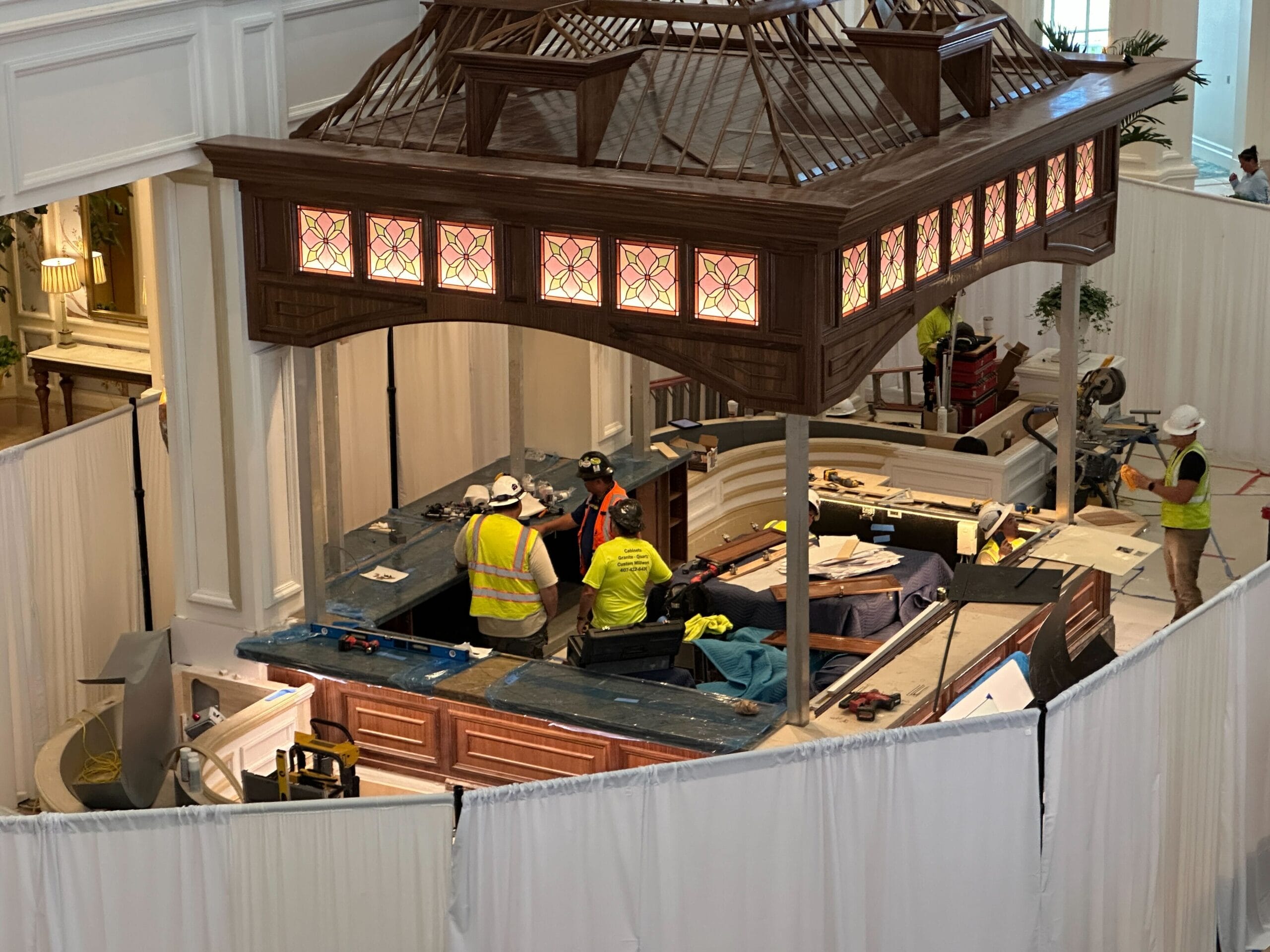 Workers in safety vests install countertops at the Grand Floridian Birdcage Bar beneath a gazebo, new lights glowing overhead.