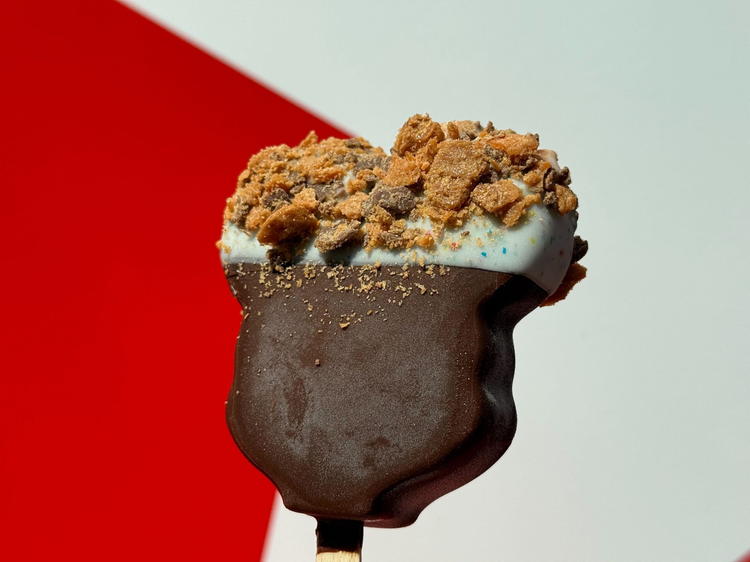 A chocolate-dipped ice cream bar with cookie crumbles, held up against a red and white backdrop typical of Main Street, U.S.A.