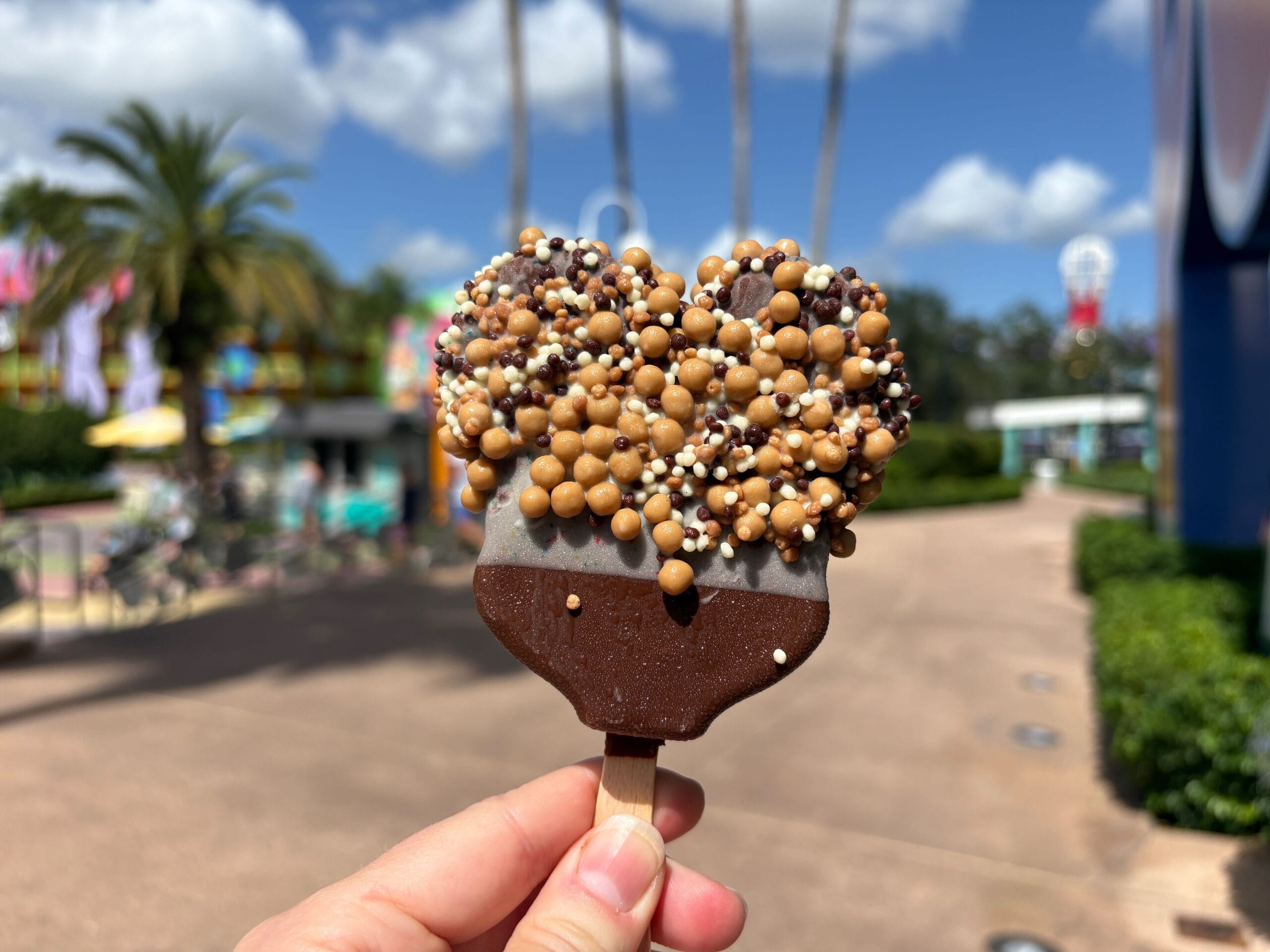 A hand holds a heart-shaped chocolate ice cream bar with candy bits at a Disney park on a sunny day, with festive vibes.