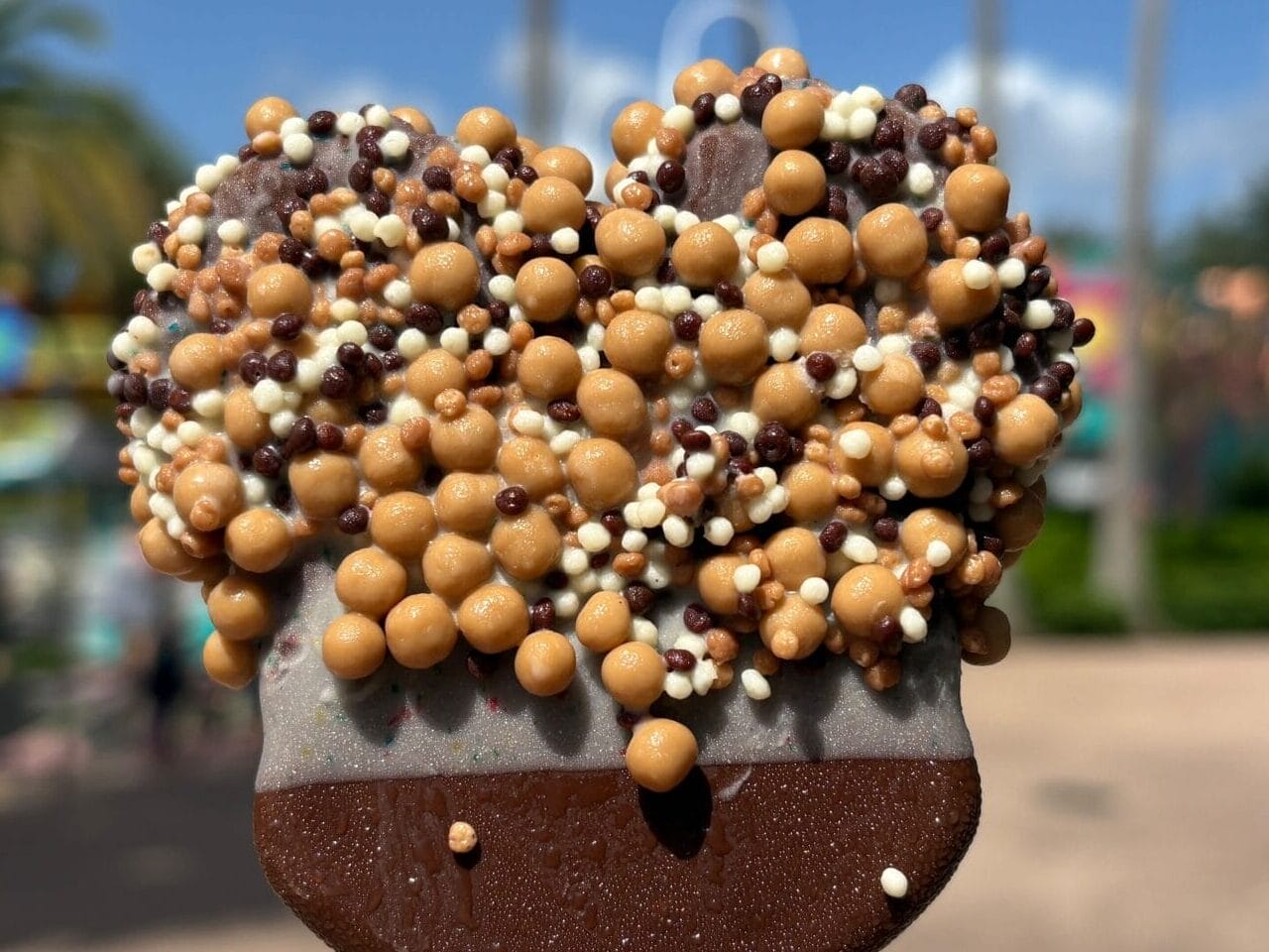 A hand holds a Mickey Mouse-shaped chocolate ice cream bar with crunchy caramel balls, enjoyed in the sunny atmosphere of a Disney park.