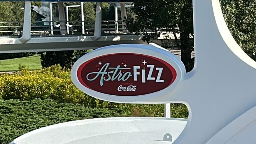Red oval "Astro FiZZ Coca-Cola" sign on a white structure, greenery behind—evoking retro Tomorrowland vibes like in Disneyland Park.