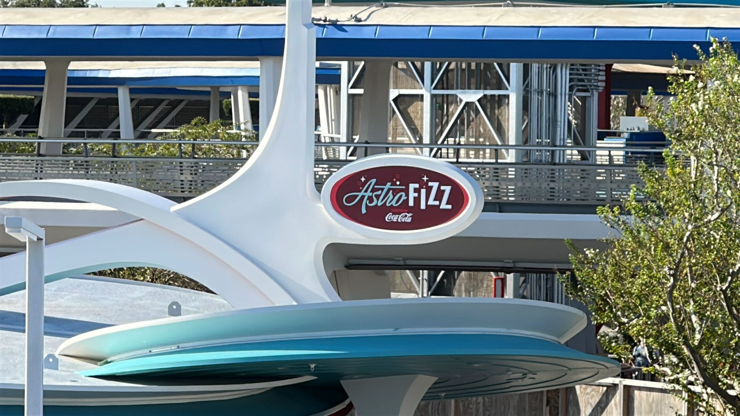 A sign reads “Astro Fizz Coca-Cola” on a retro-futuristic Magic Kingdom building with blue and white accents—classic Tomorrowland style.