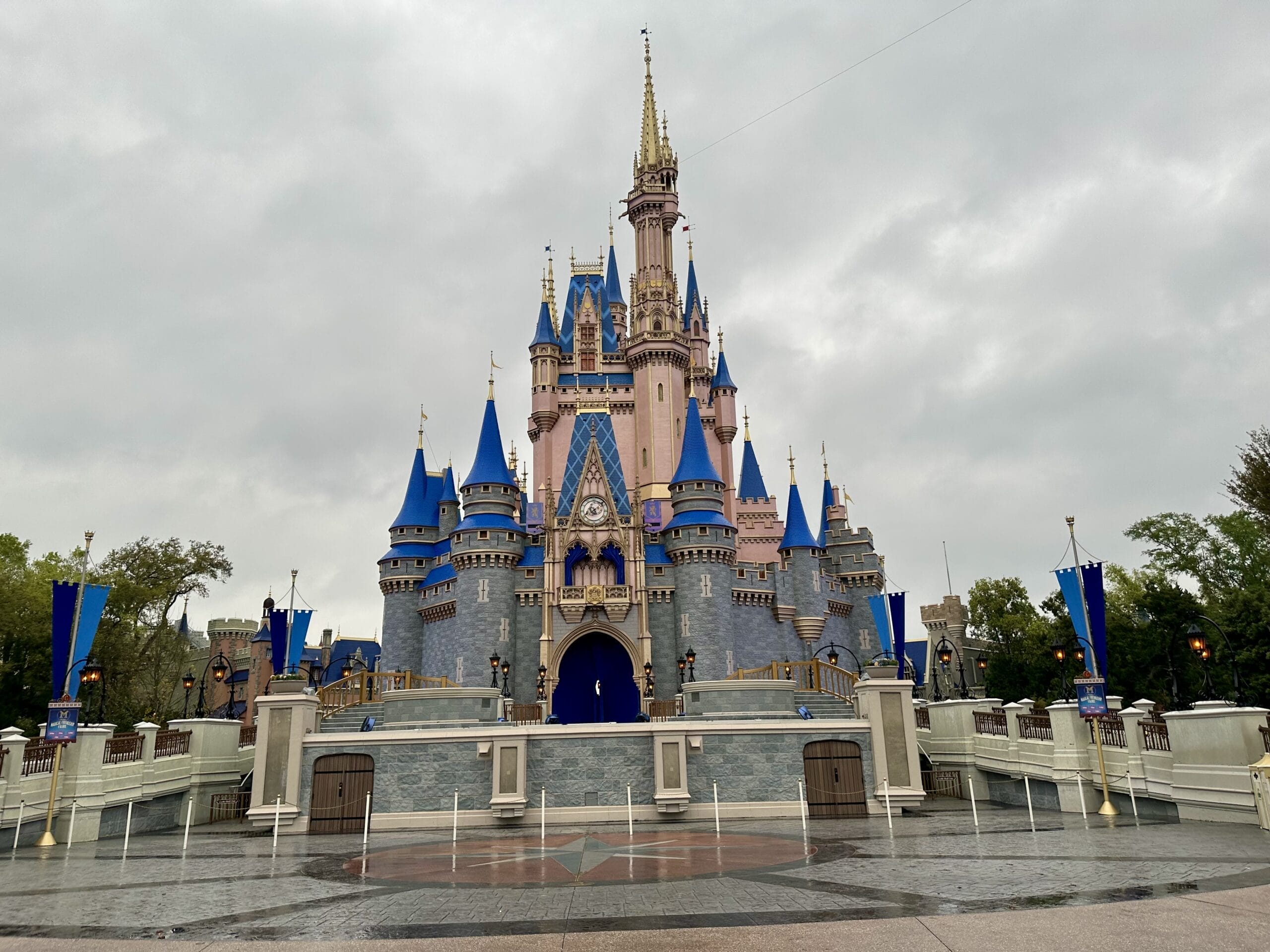 Cinderella Castle at Walt Disney World, noted for its blue and gold details, stands empty under clouds—taller than Disneyland’s castle.