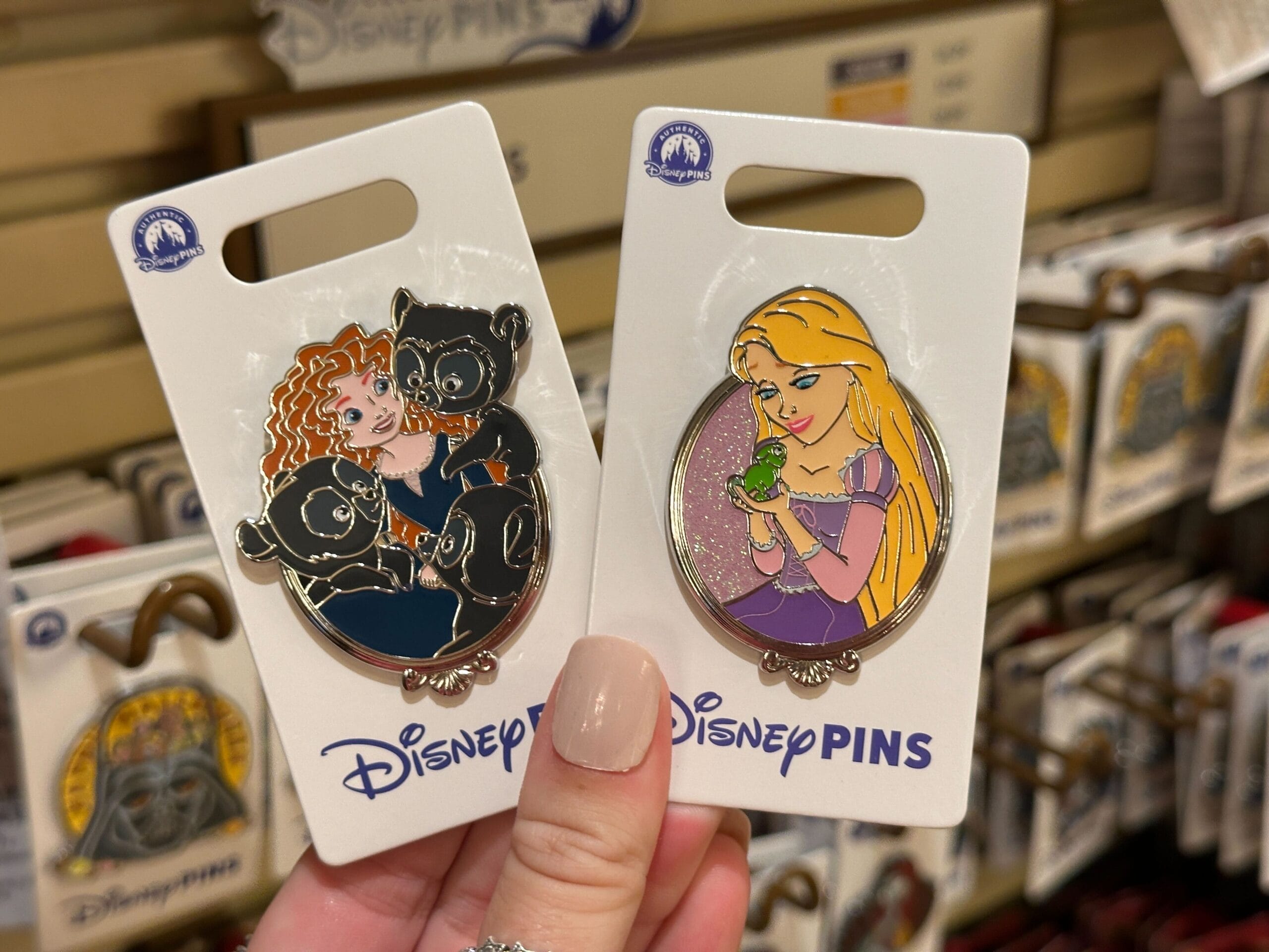 Hand displays Walt Disney World Sidekicks Pins of Merida with bear cubs and Rapunzel holding Pascal—no castle shown to compare.