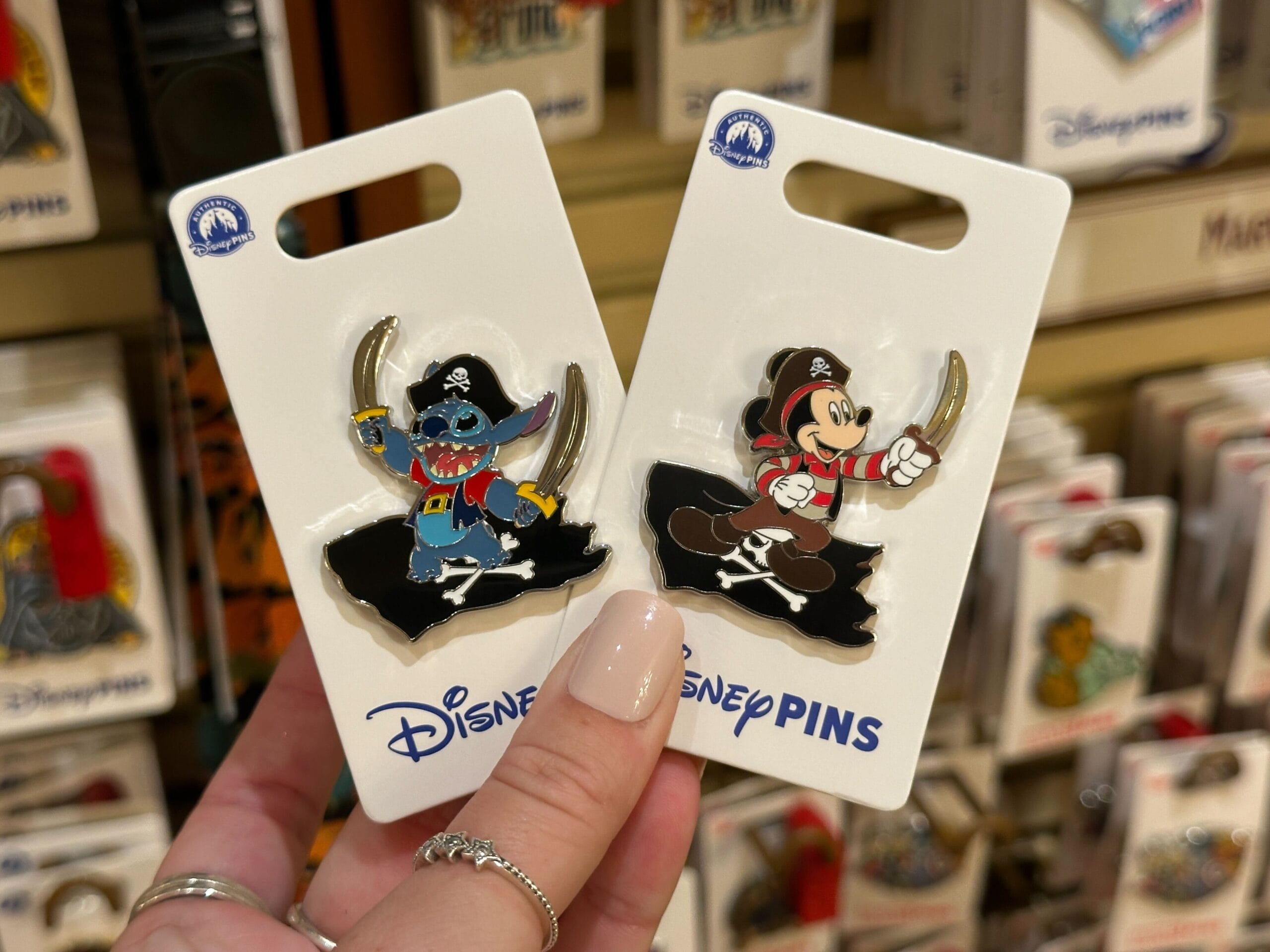 Hand holding Stitch Pirate Pins and Minnie pirate pins in packaging, seen at a Disney World store near Cinderella Castle.