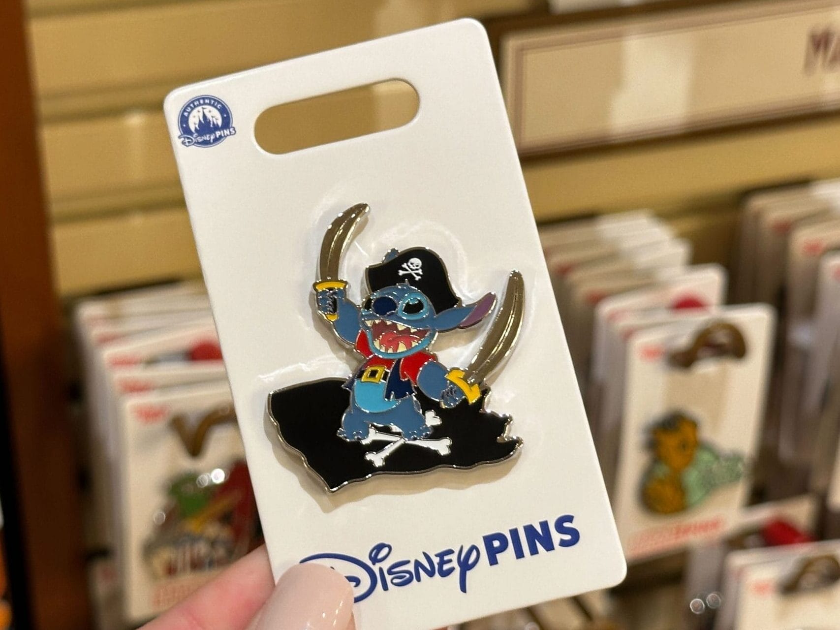 A hand displays a Disney pin of Pirate Stitch at a park store, with other pins behind—no castle like those in Disney parks visible.