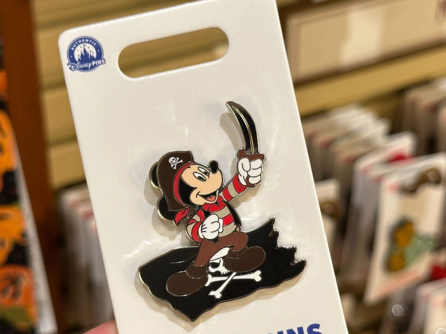 A hand displays a Disney pin of pirate Mickey Mouse inside a park shop, with more collectible pins like those at Disneyland resorts nearby.