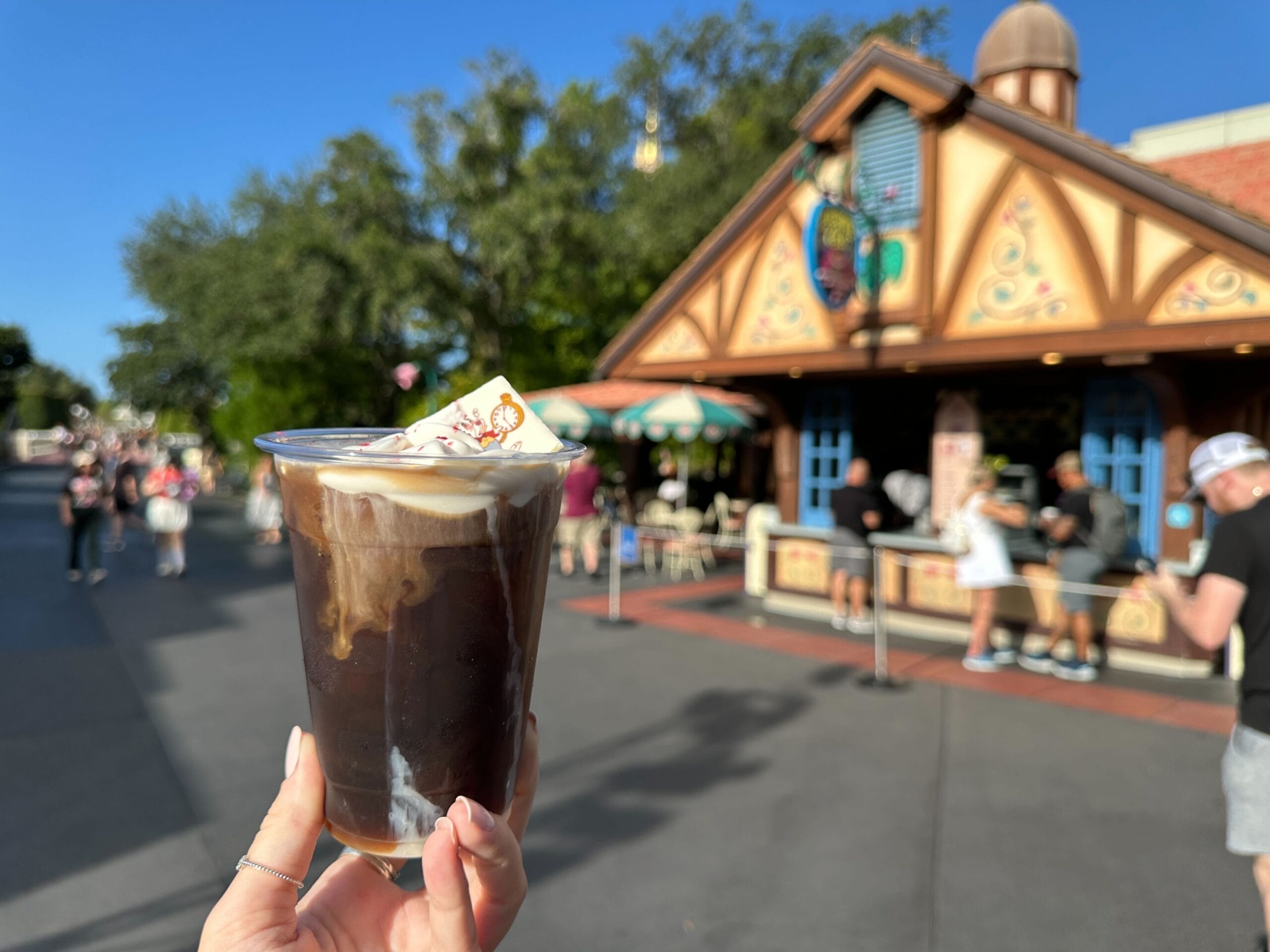 A hand holds a cold brew coffee beverage with a White Rabbit chocolate piece with Fantasyland in Magic Kingdom in the background.