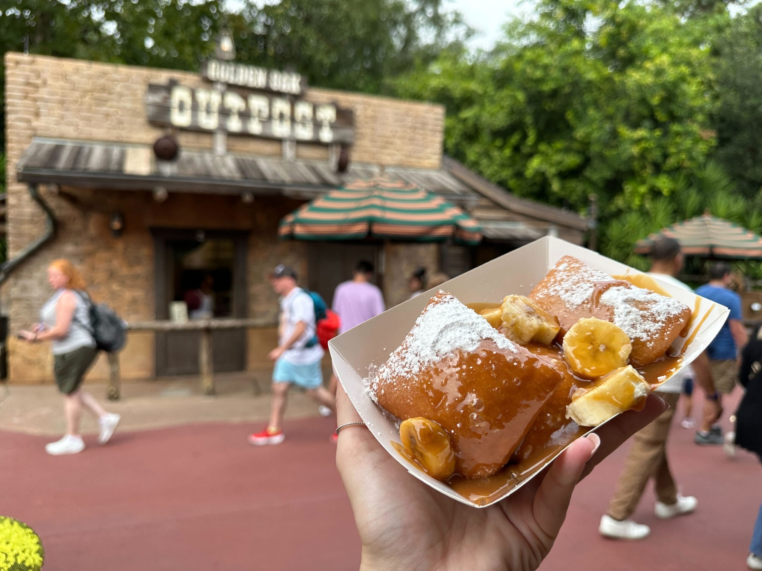 A hand holds banana-caramel beignets at Magic Kingdom's Golden Oak Outpost, no castle in view as guests walk past.
