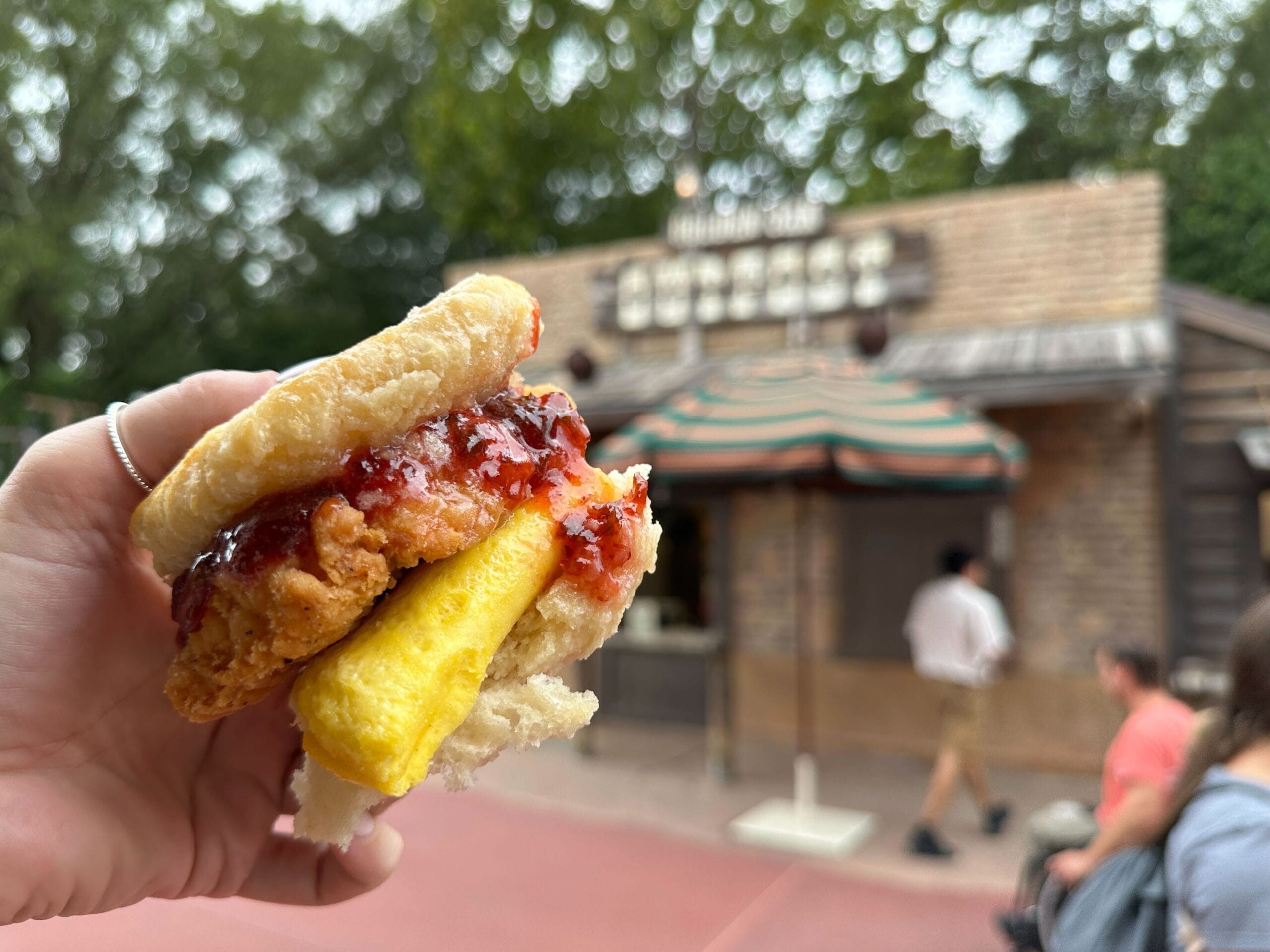 At Magic Kingdom’s Golden Oak Outpost, a hand holds a biscuit breakfast sandwich stacked with egg, fried chicken, and jelly.