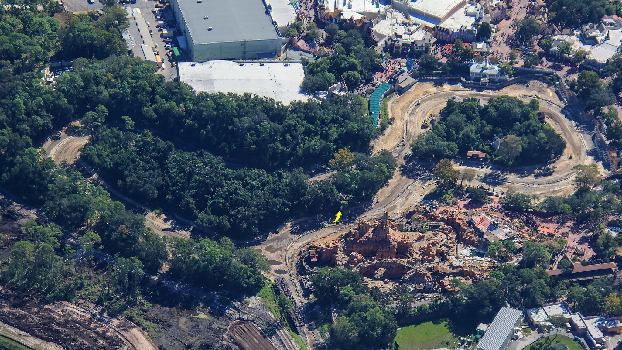 Aerial view of a Disney park with winding dirt paths, dense trees, and Rivers of America’s rocky structure being built, no castle visible.