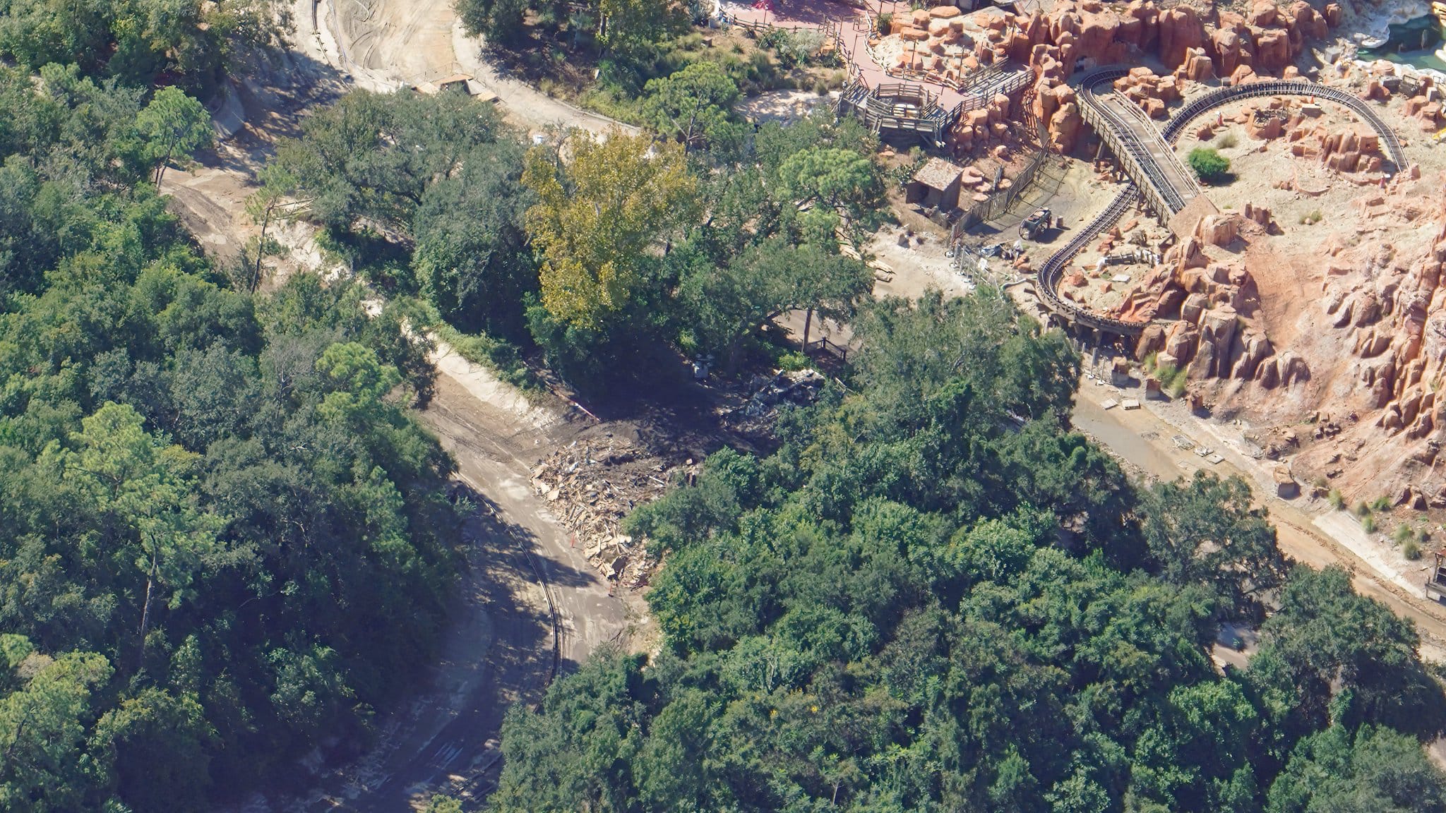 Aerial view of a tree-lined dirt path near a roller coaster and rocky landscape in a Disney park, reminiscent of Frontierland.