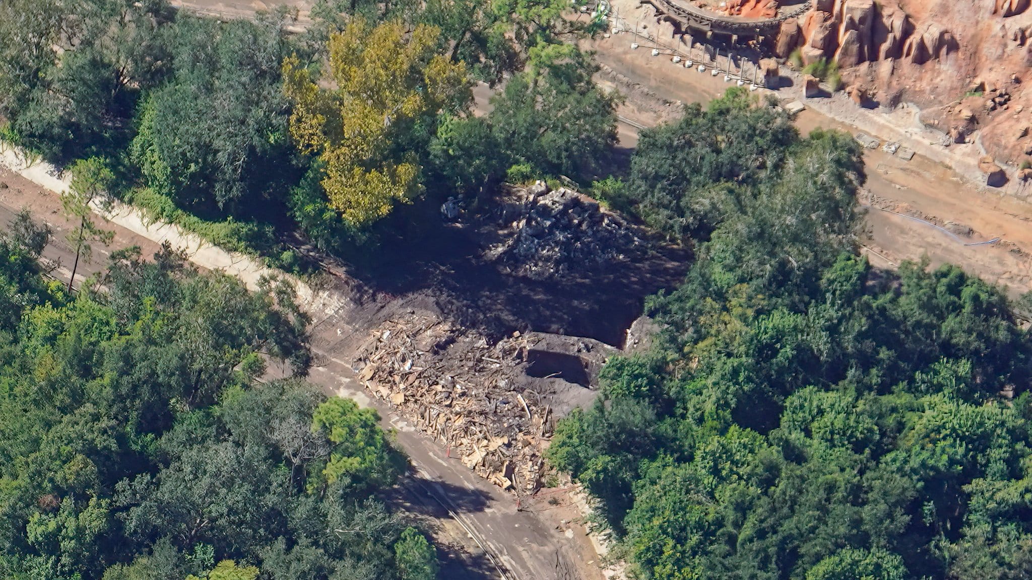 Aerial view of a demolition site with debris piles amid dense greenery, resembling wooded areas near Disney parks but no castle present.