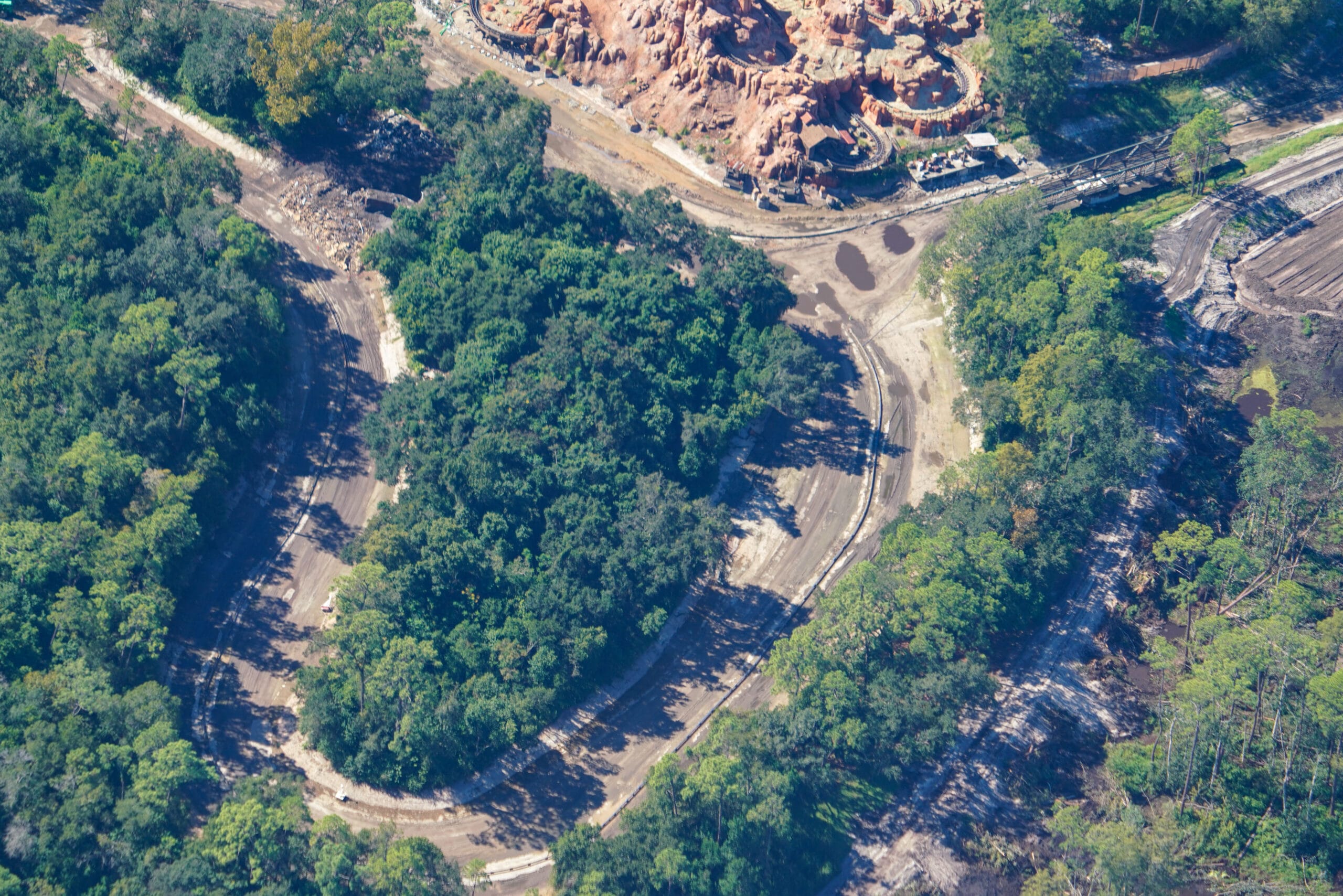 Aerial view of a winding dirt path through lush forest by rugged terrain, styled like Disneyland's Rivers of America landscaping.