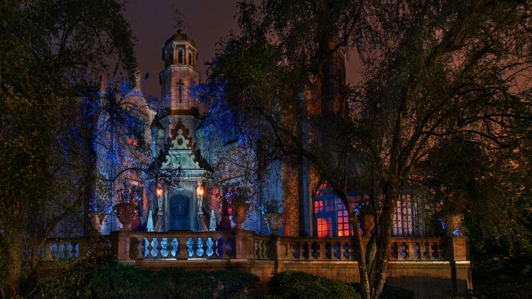 The Haunted Mansion at night, illuminated by blue and red lights, partially obscured by trees—unlike any Disney castle design.