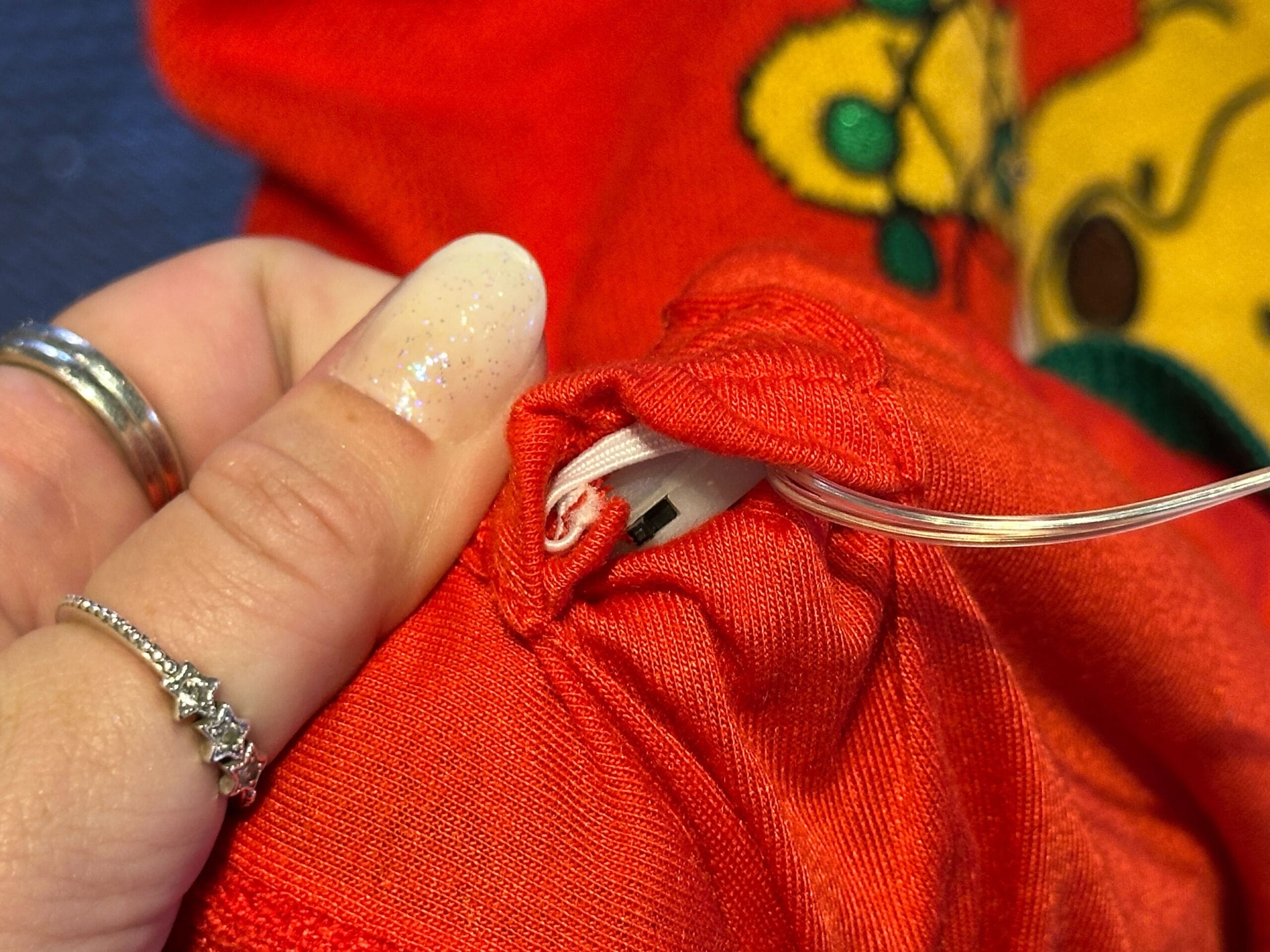 A hand holds red fabric from a Light-Up Winnie the Pooh sweater at Disney, showing a white device through an open seam.