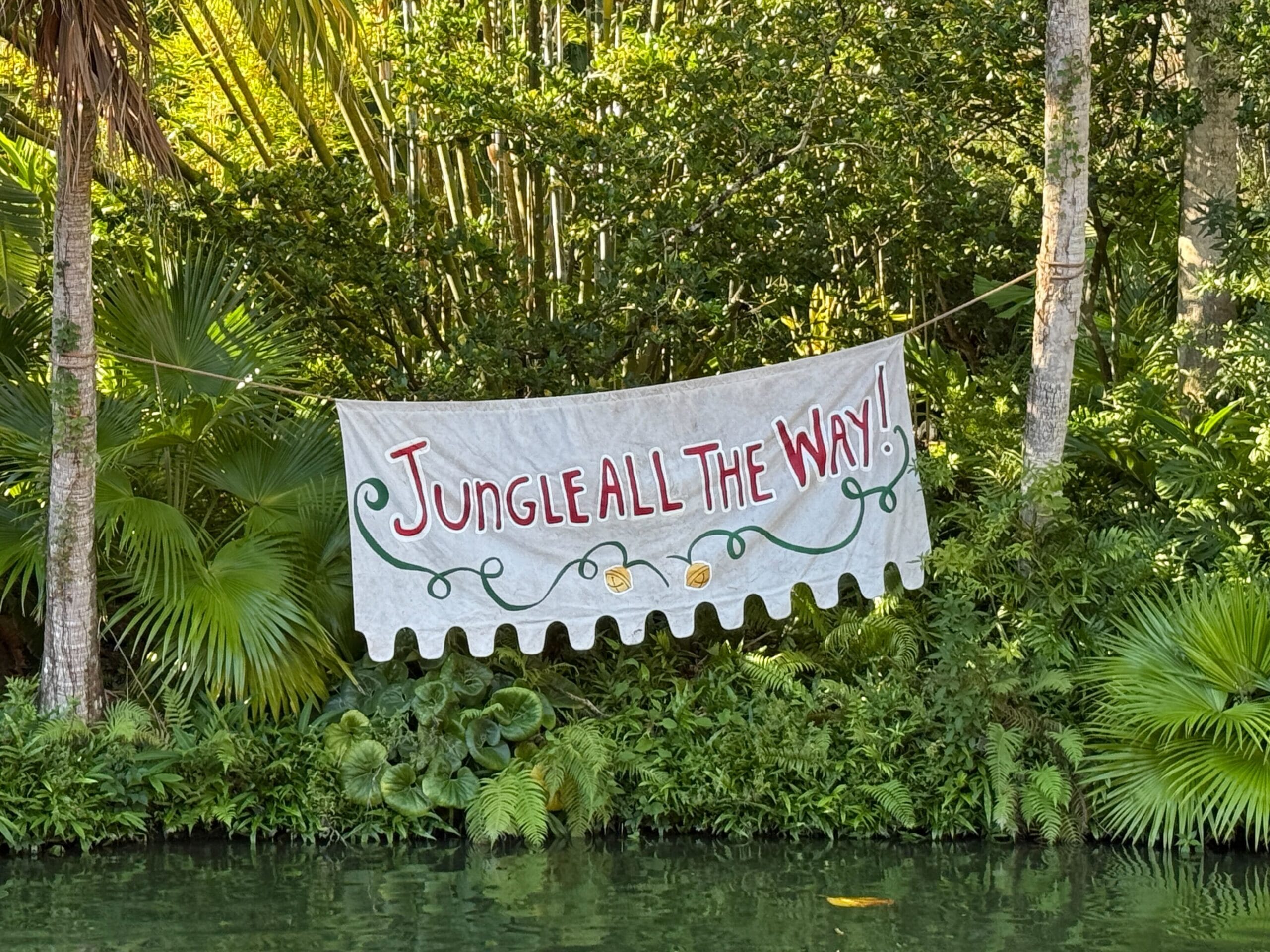 A “Jungle All The Way!” banner adds Jingle Cruise festive flair to tropical foliage and water at Magic Kingdom