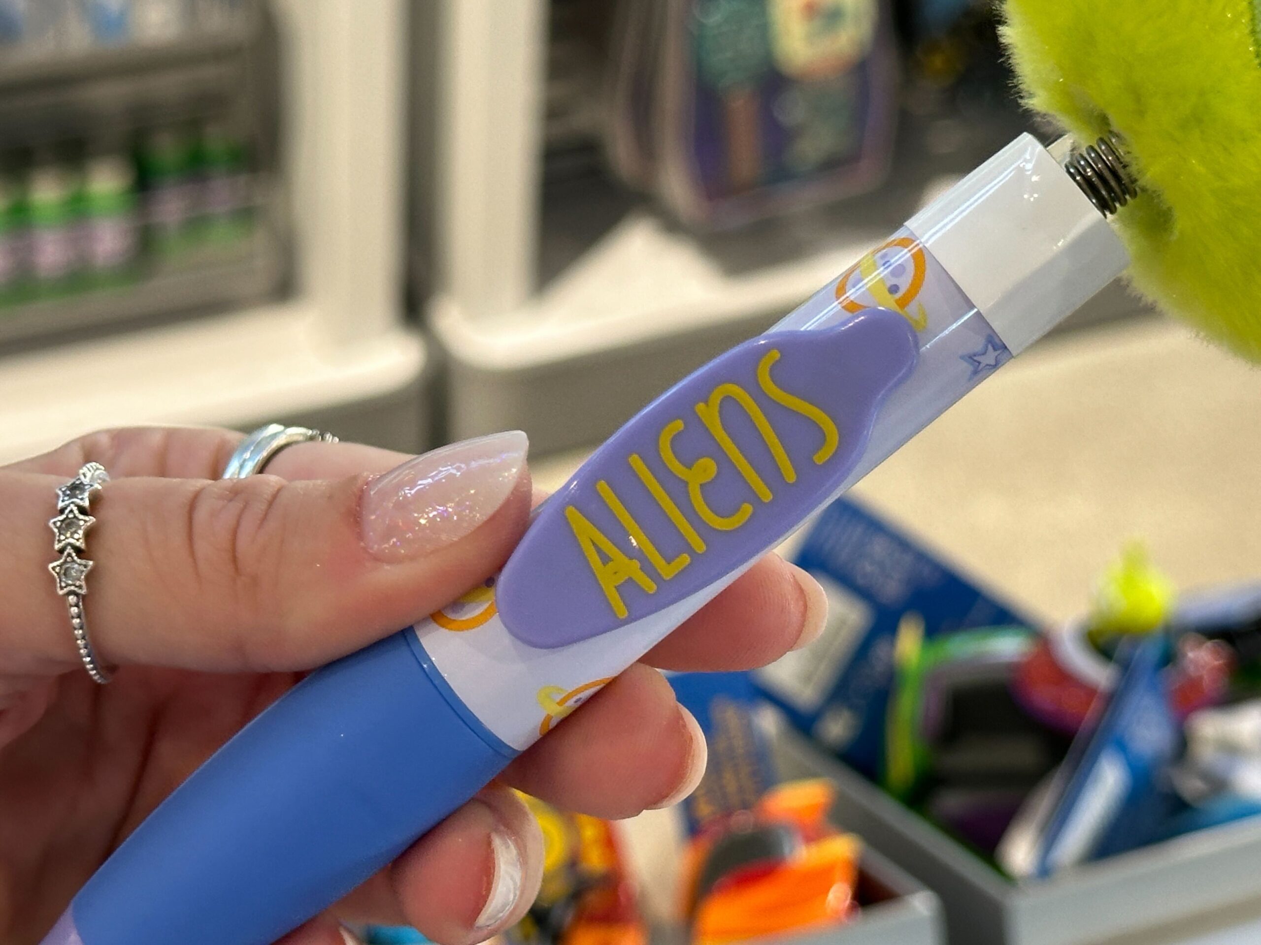 A hand holds a blue Toy Story 30th Anniversary pen with a fuzzy top and "ALIENS" printed on it, sold at Disney parks worldwide.