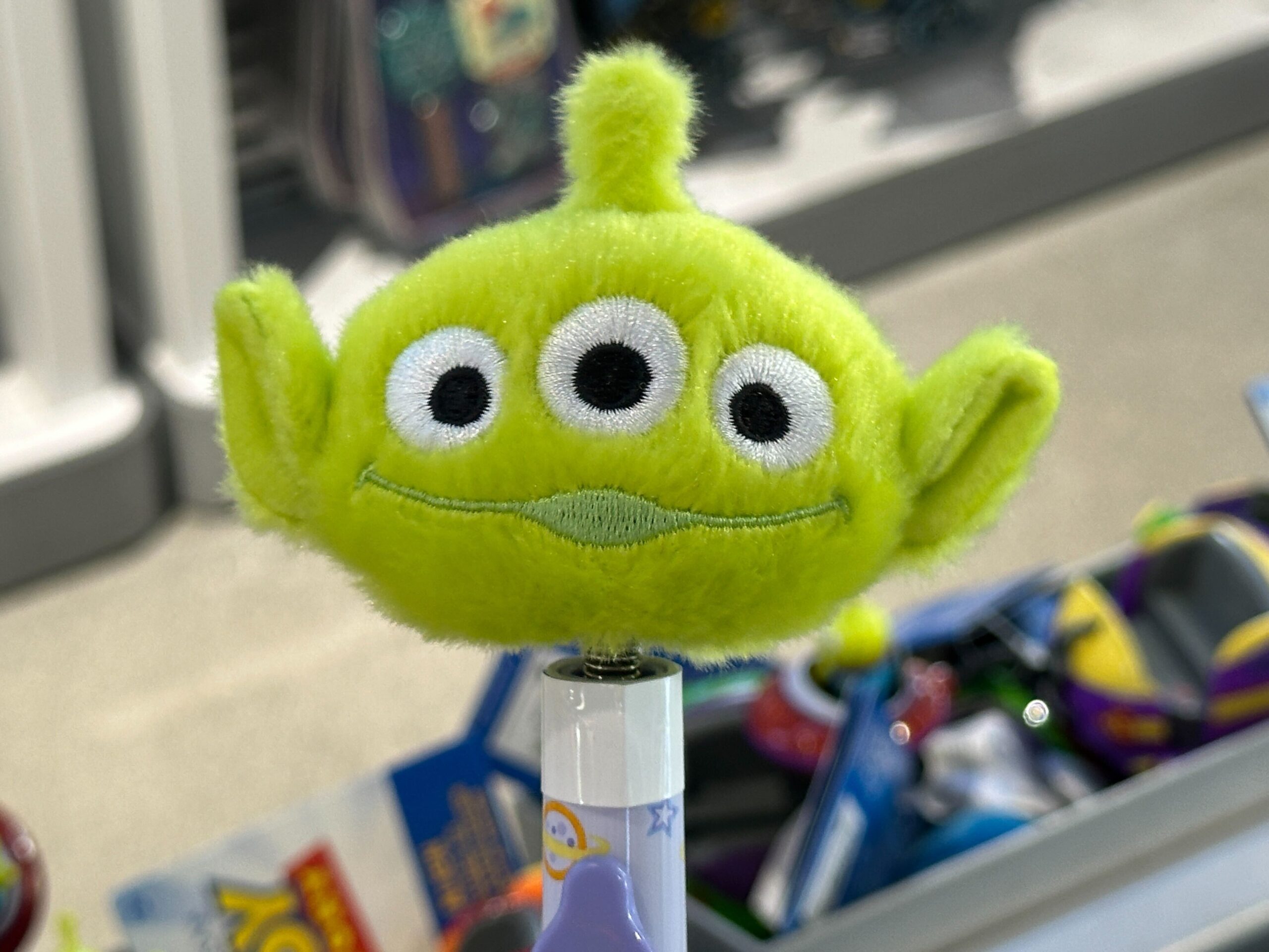 A plush green alien with three eyes and pointy ears sits atop a pen at a Disney World shop, surrounded by park-themed displays.