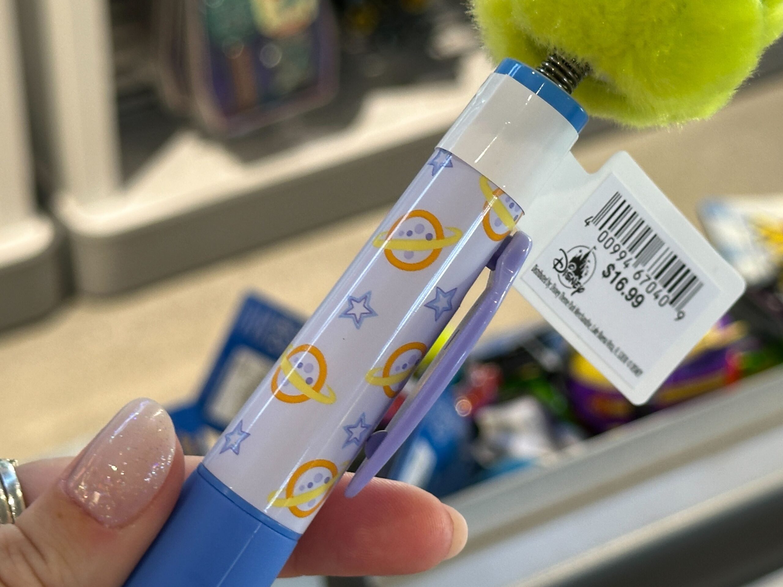 A hand holds a blue pen with yellow planet and star designs, marked as Toy Story 30th Anniversary for $16.99, found at Disney Parks.