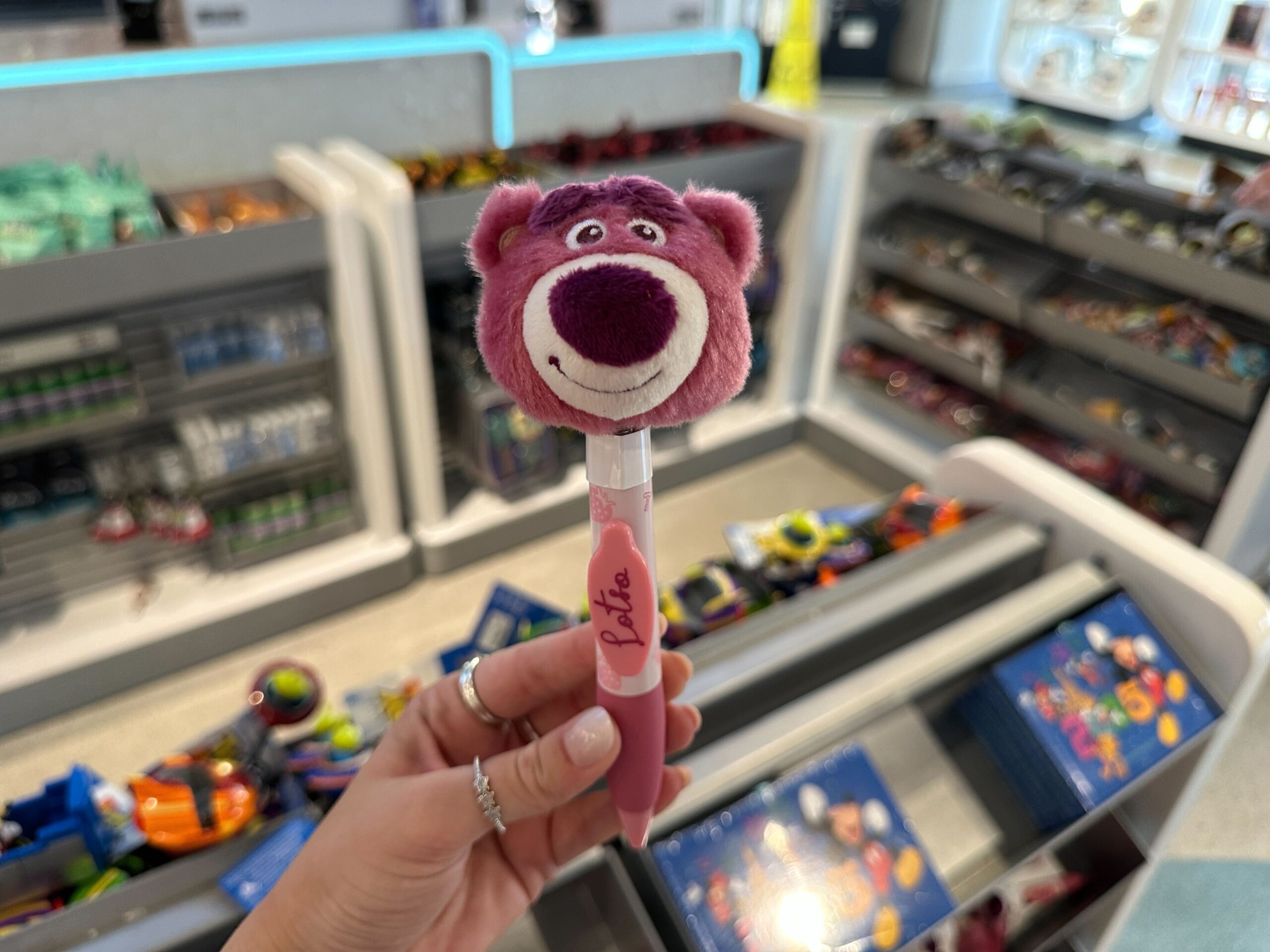 A hand holds a plush Lotso bear pen in a Disney park store aisle, Toy Story 30th Anniversary decor visible in the background.