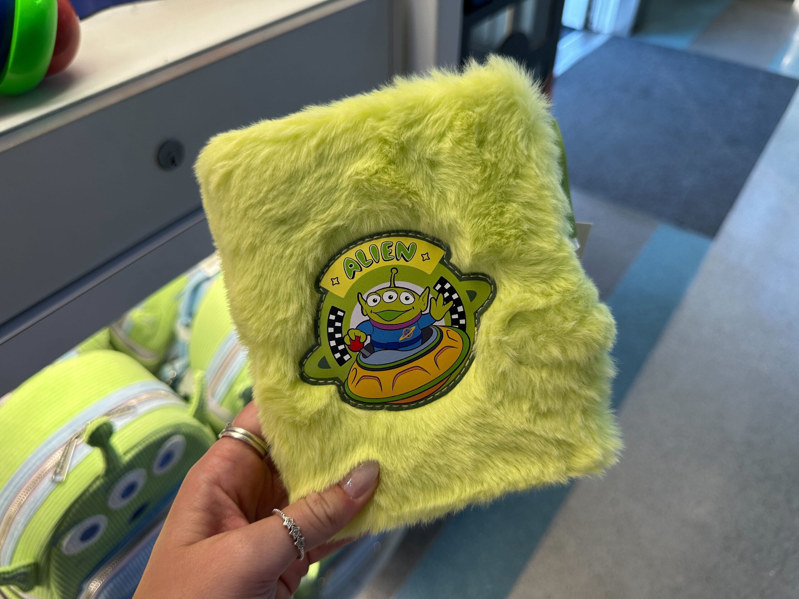 A hand holds a fuzzy green Alien notebook with a Toy Story alien, sold for the 30th Anniversary—much like park-exclusive Disney merch.