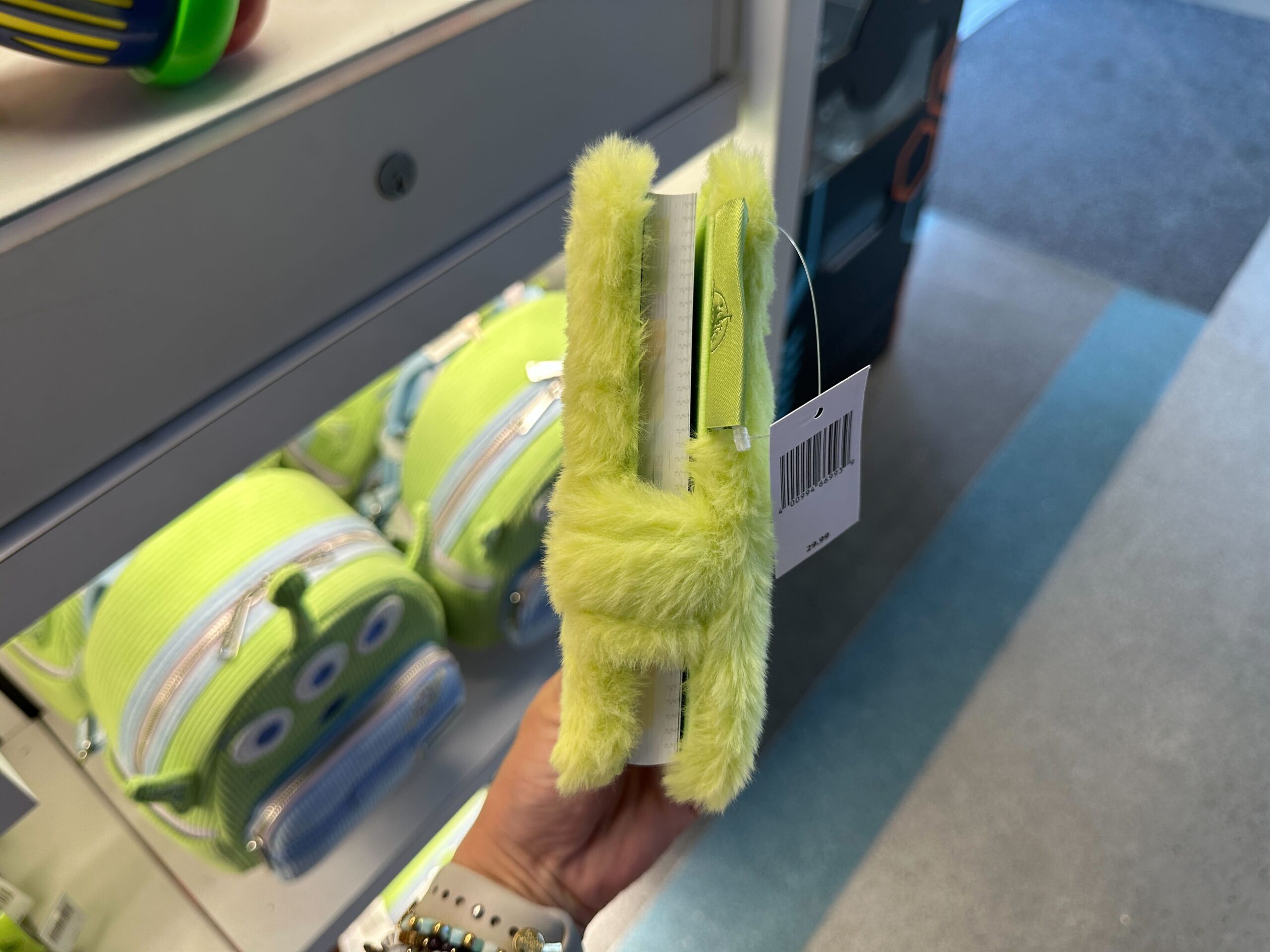 A hand holds a fuzzy green pen like a clothespin in a Disney store, Toy Story 30th Anniversary alien backpacks on display behind.