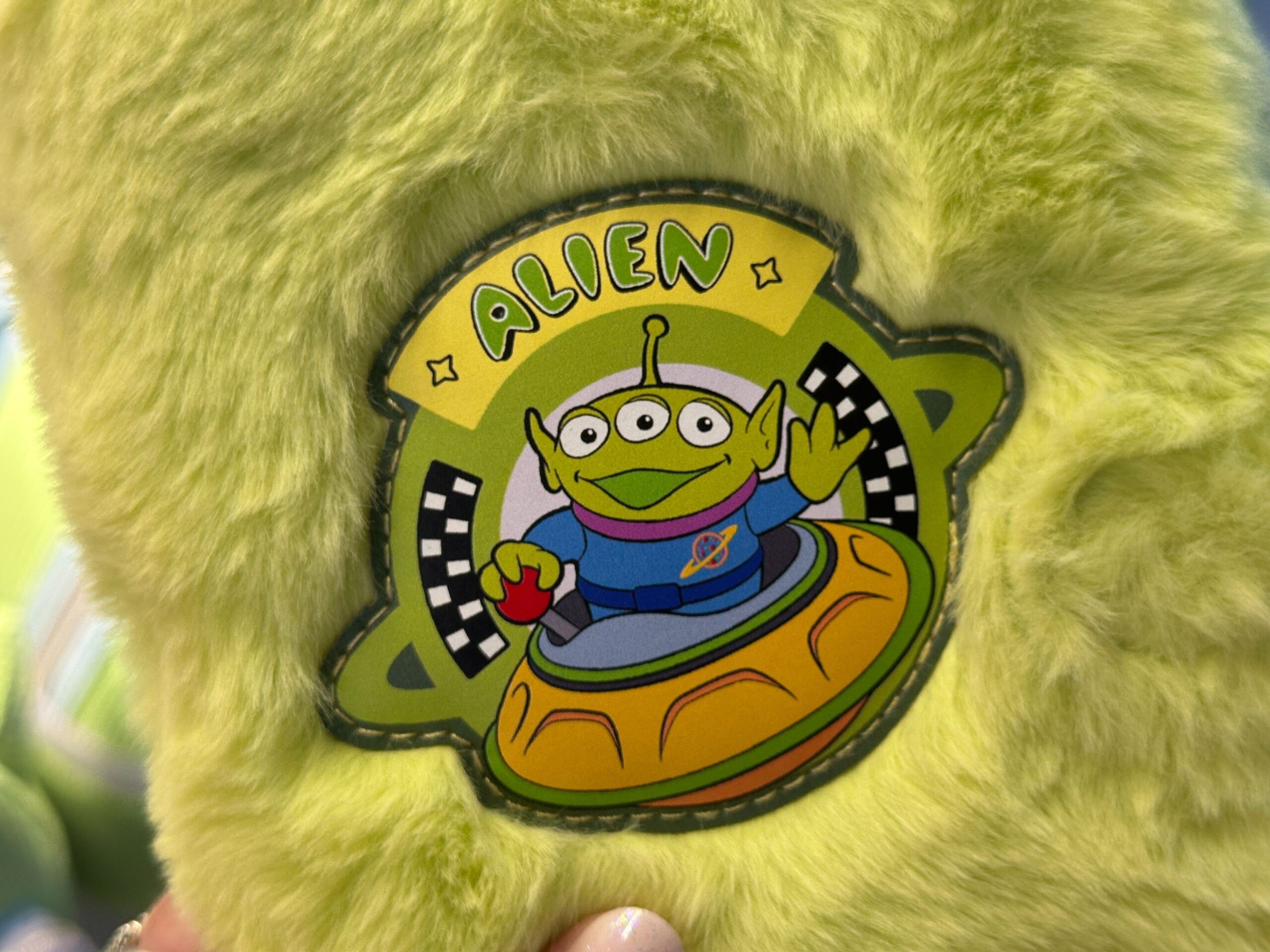 A green plush toy with a patch featuring a Toy Story 30th Anniversary alien in a spaceship, "ALIEN" above—seen at Disney Parks.