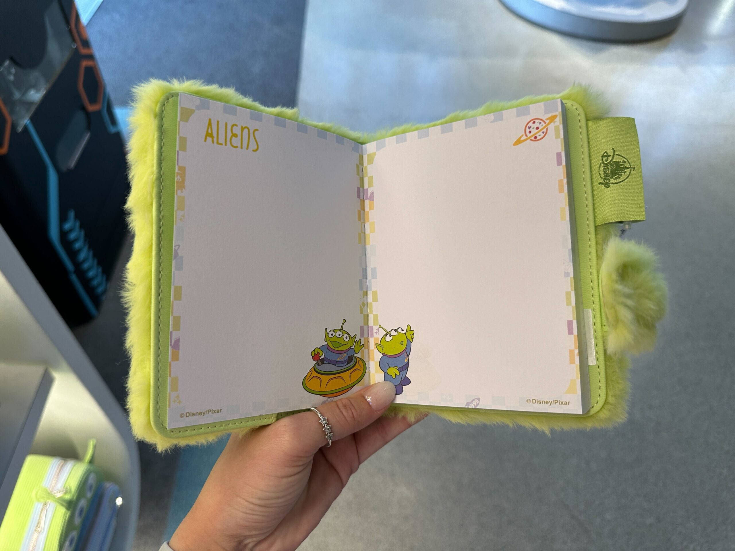 A hand displays a fuzzy green "Aliens" Toy Story 30th Anniversary notebook, echoing the playful vibe of Disney park merchandise.