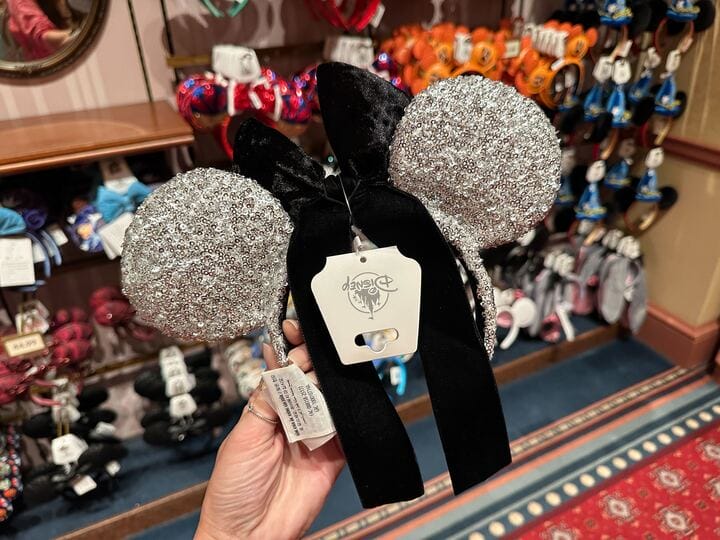 Hand holding sparkly silver Minnie Mouse ears with black velvet bow in a Disney park shop, other ear headbands on display nearby.