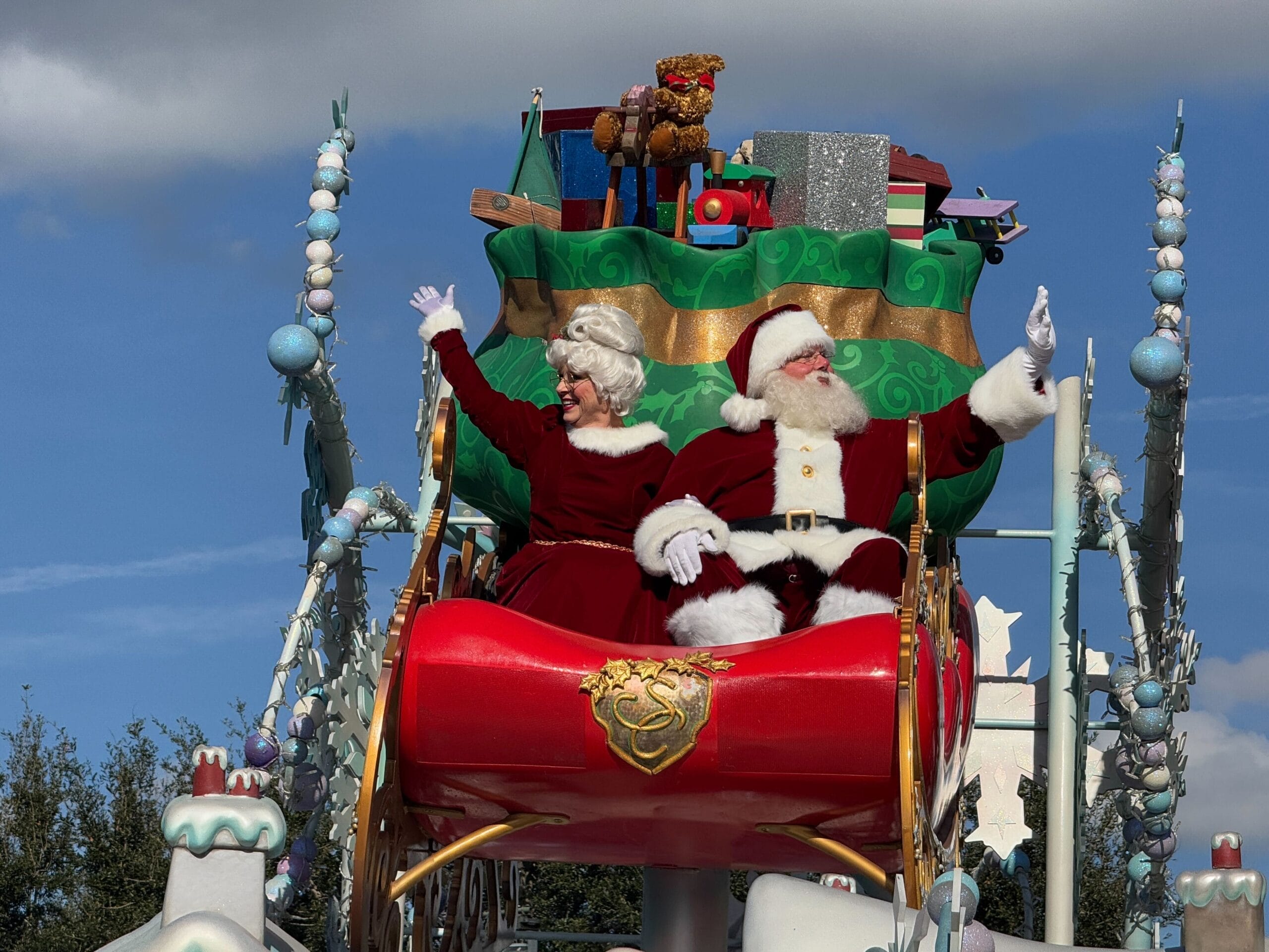 Santa and Mrs. Claus wave from a red sleigh in a Disney park parade, surrounded by Park Hopper magic and joyful holiday spirit.